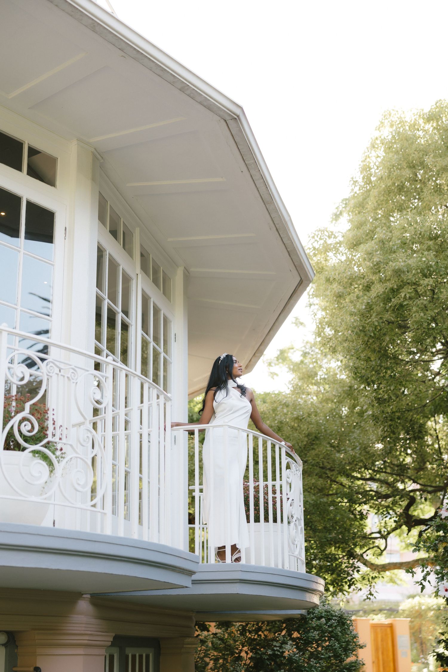 Bride in white dress poses on classical white balcony surrounded by greenery.
