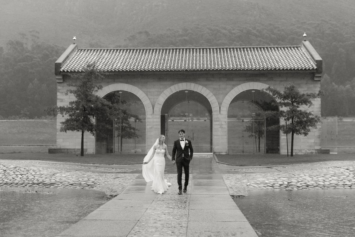 Black and white photo of newlyweds walking toward ornate arched building entrance.