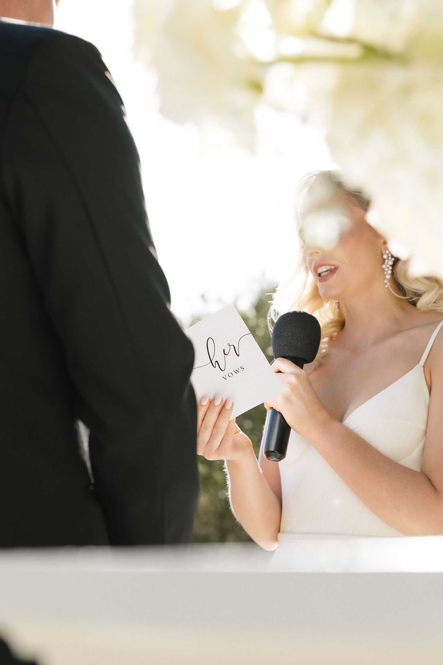 Bride reading her vows from a card into a microphone during an outdoor wedding ceremony.