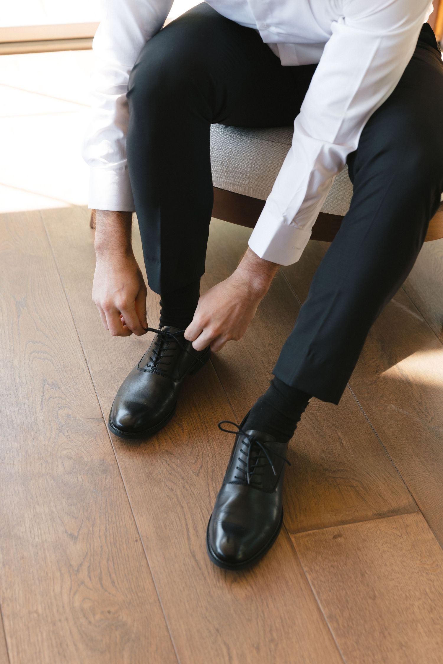 Close-up of groom tying classic black leather dress shoes on wooden floor.