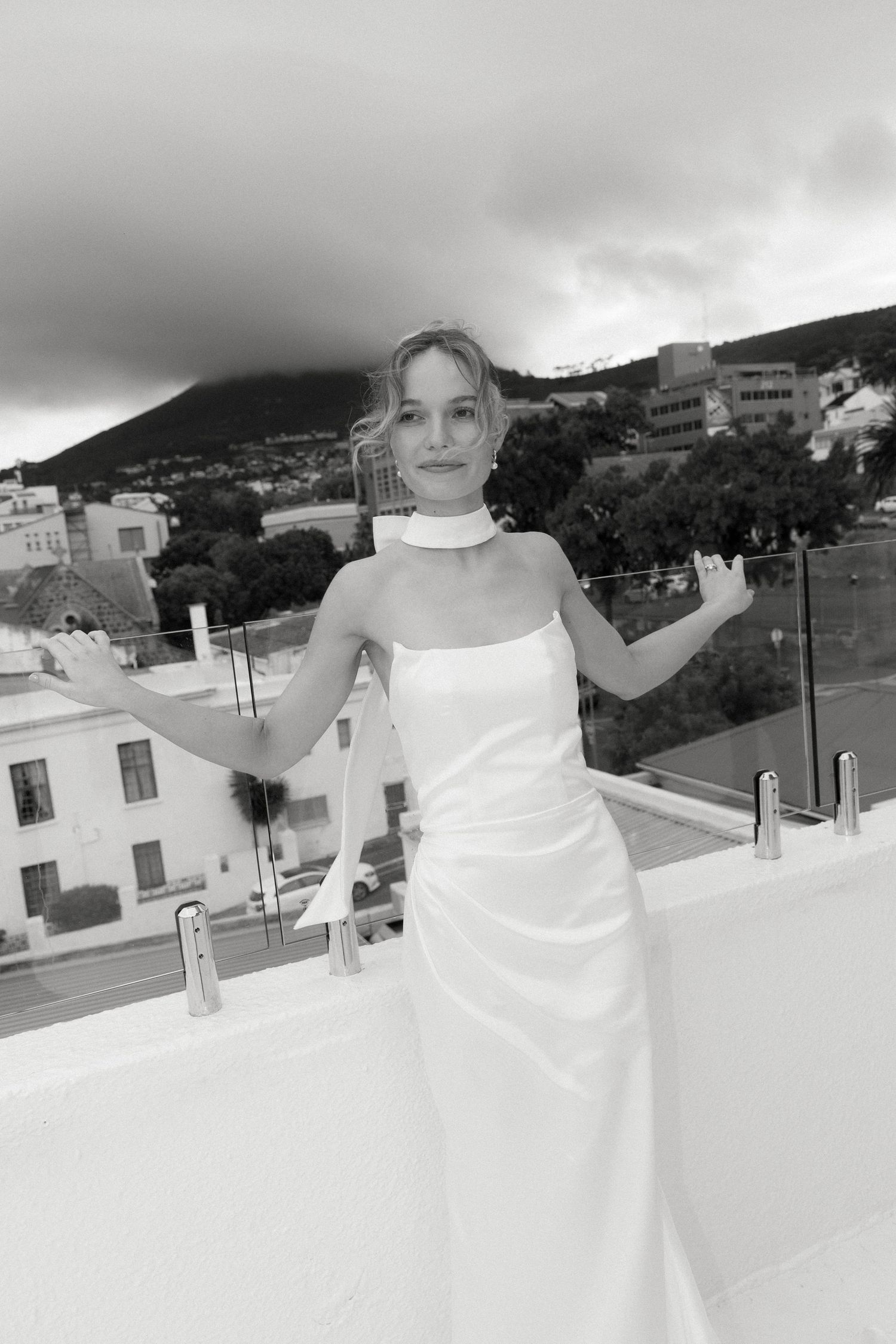 A bride poses playfully on a rooftop terrace against a dramatic cloudy mountain backdrop.