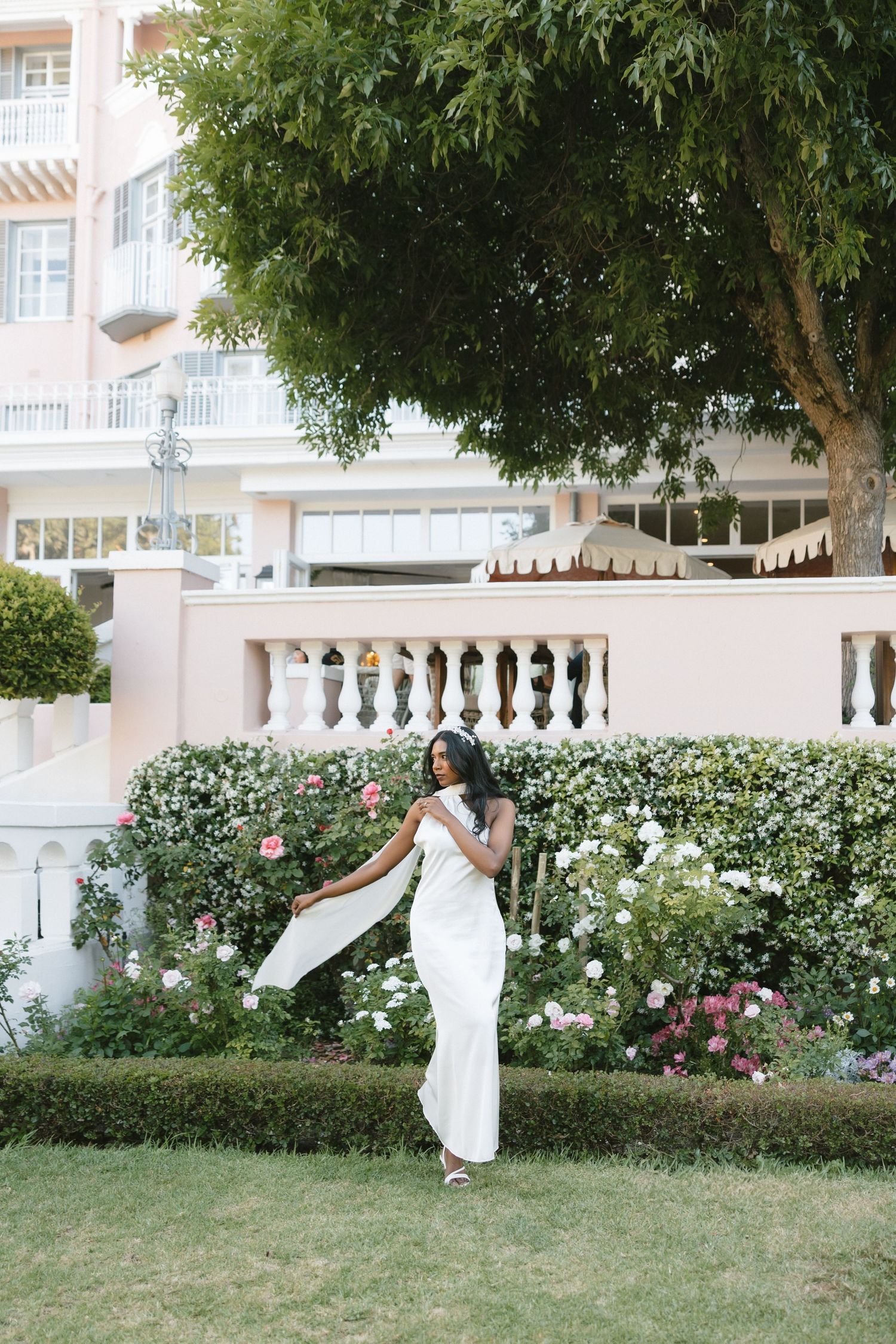 Bride in flowing white dress twirling in garden with classical architecture backdrop.