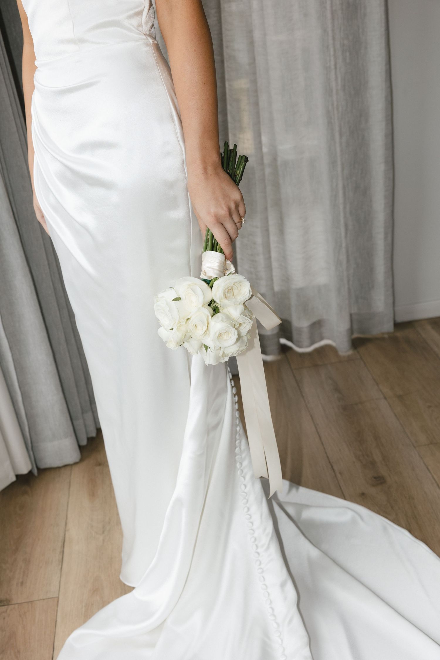 Detail of white wedding dress train and bouquet of white roses on wooden floor.