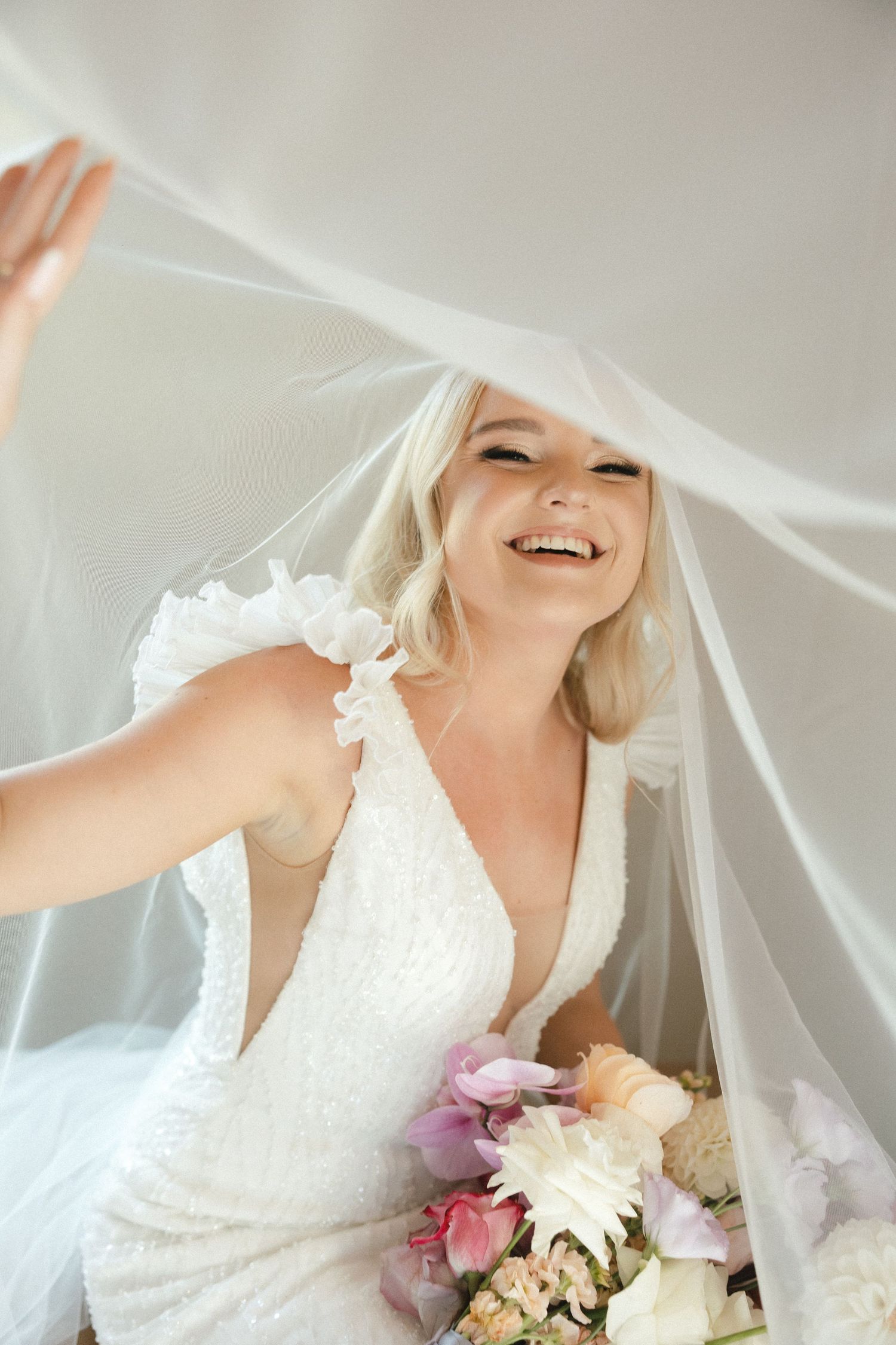 Candid moment of a bride playfully lifting her veil while holding a colorful bouquet.