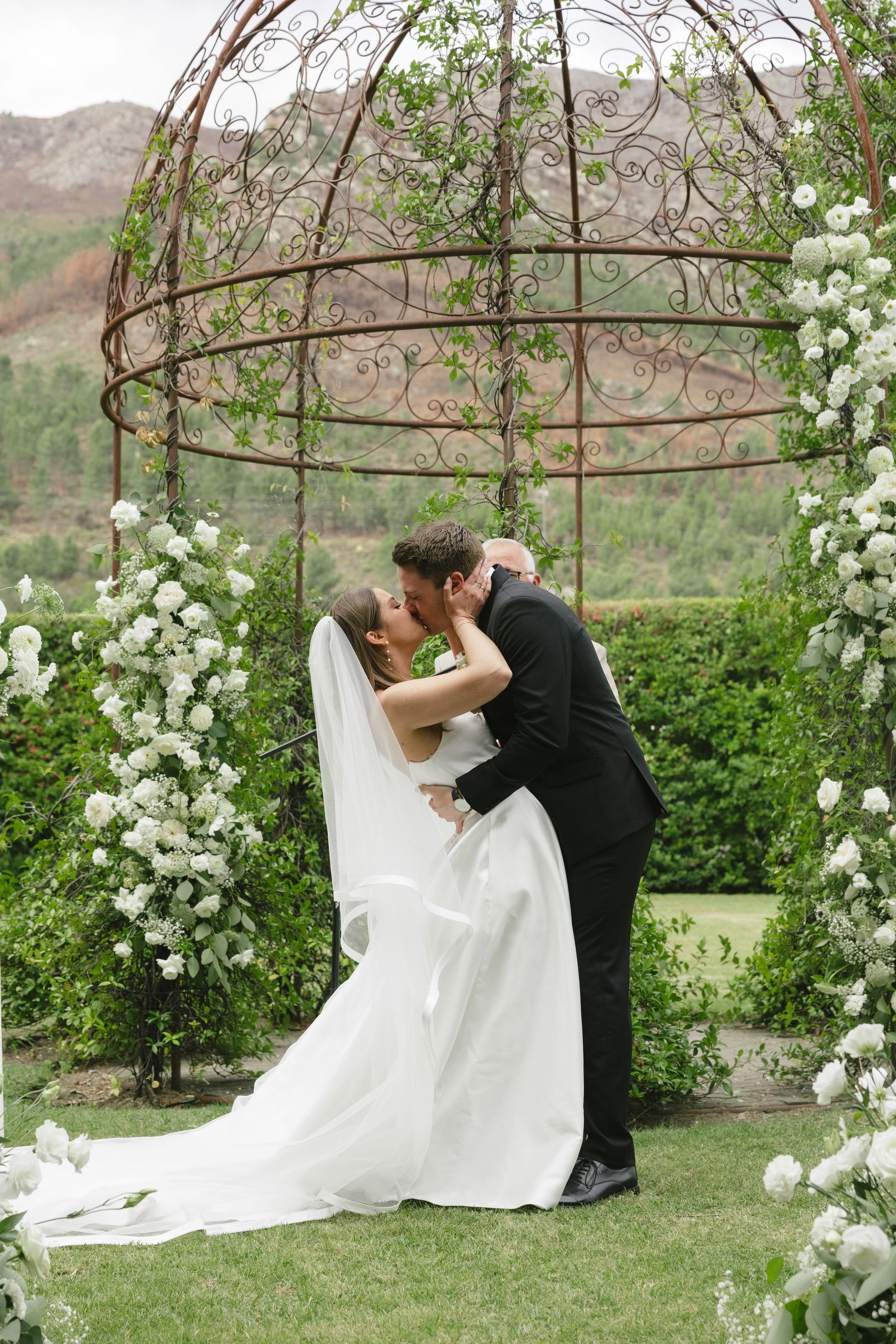 Bride and groom share first kiss under ornate iron arch adorned with white flowers in lush garden wedding ceremony.