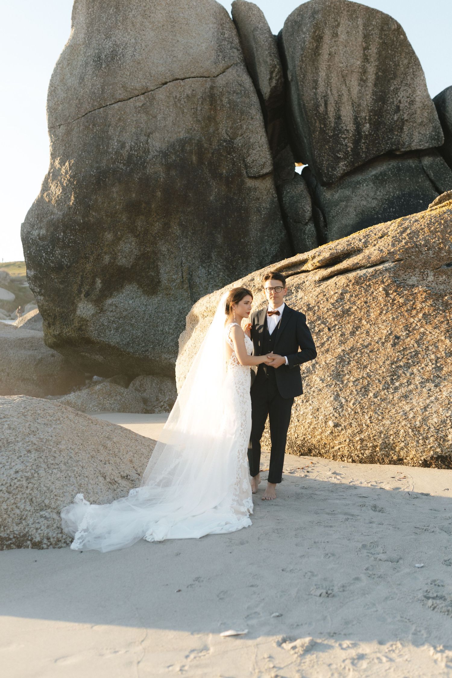 Wedding couple posing against dramatic coastal rock formations at sunset.