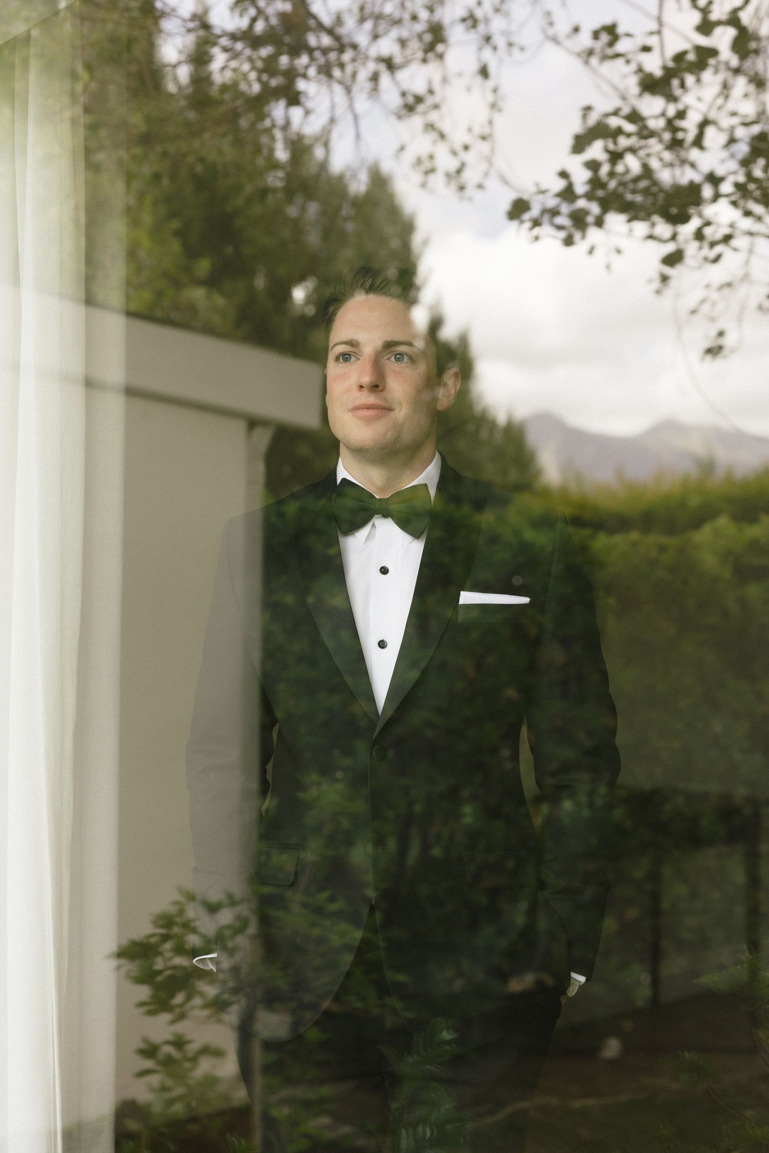 Elegant groom in black tuxedo with bow tie stands by window, lush green trees reflected in glass.