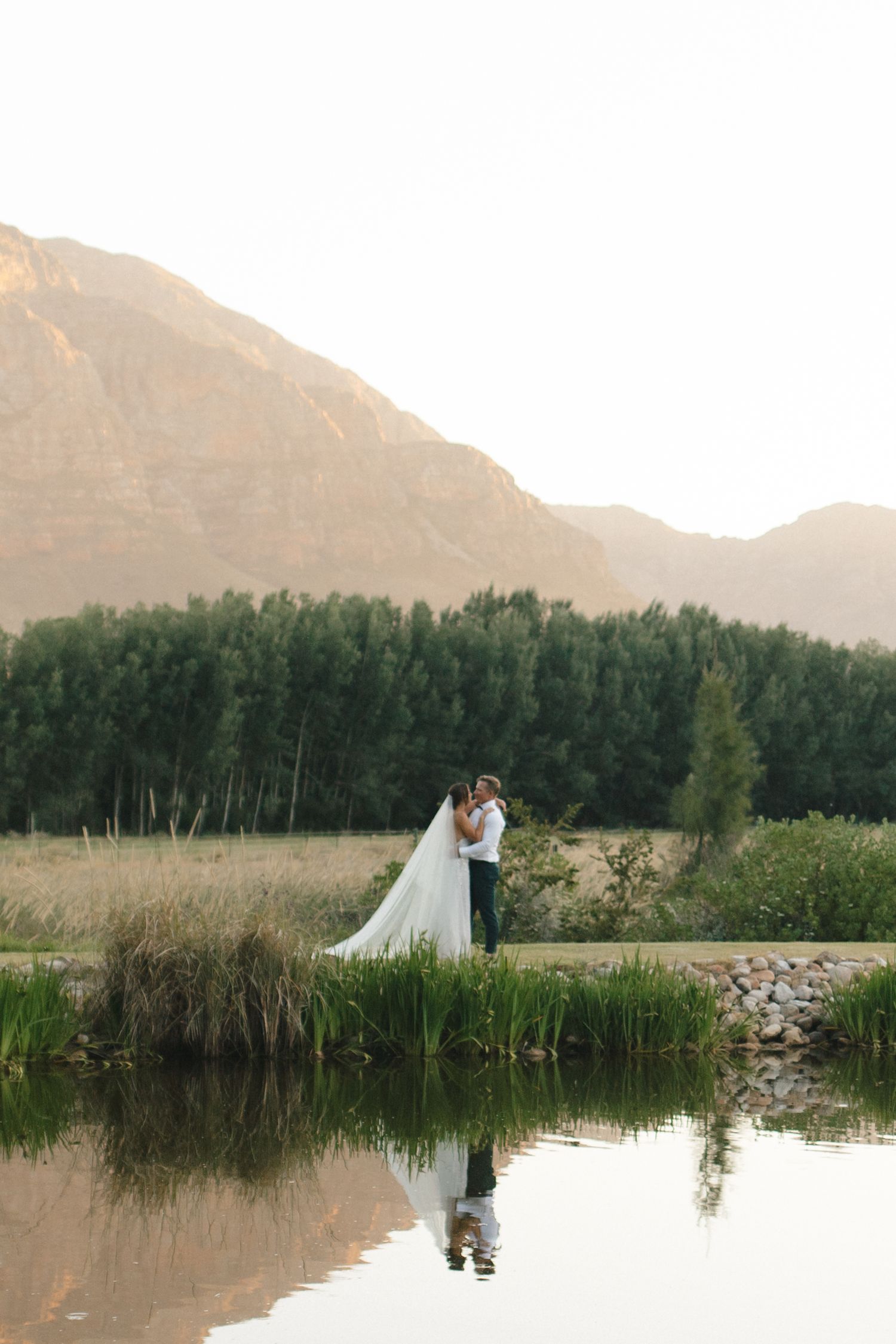 Newlyweds standing by a reflective pond with mountain landscape at sunset.