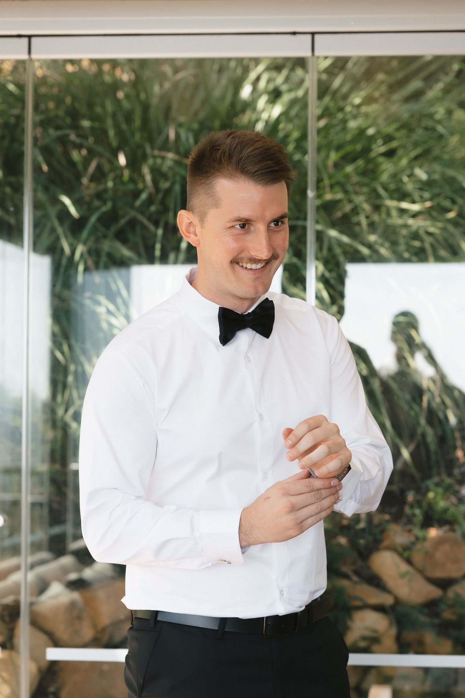 Groom in classic white dress shirt and black bow tie adjusts cufflinks by window.