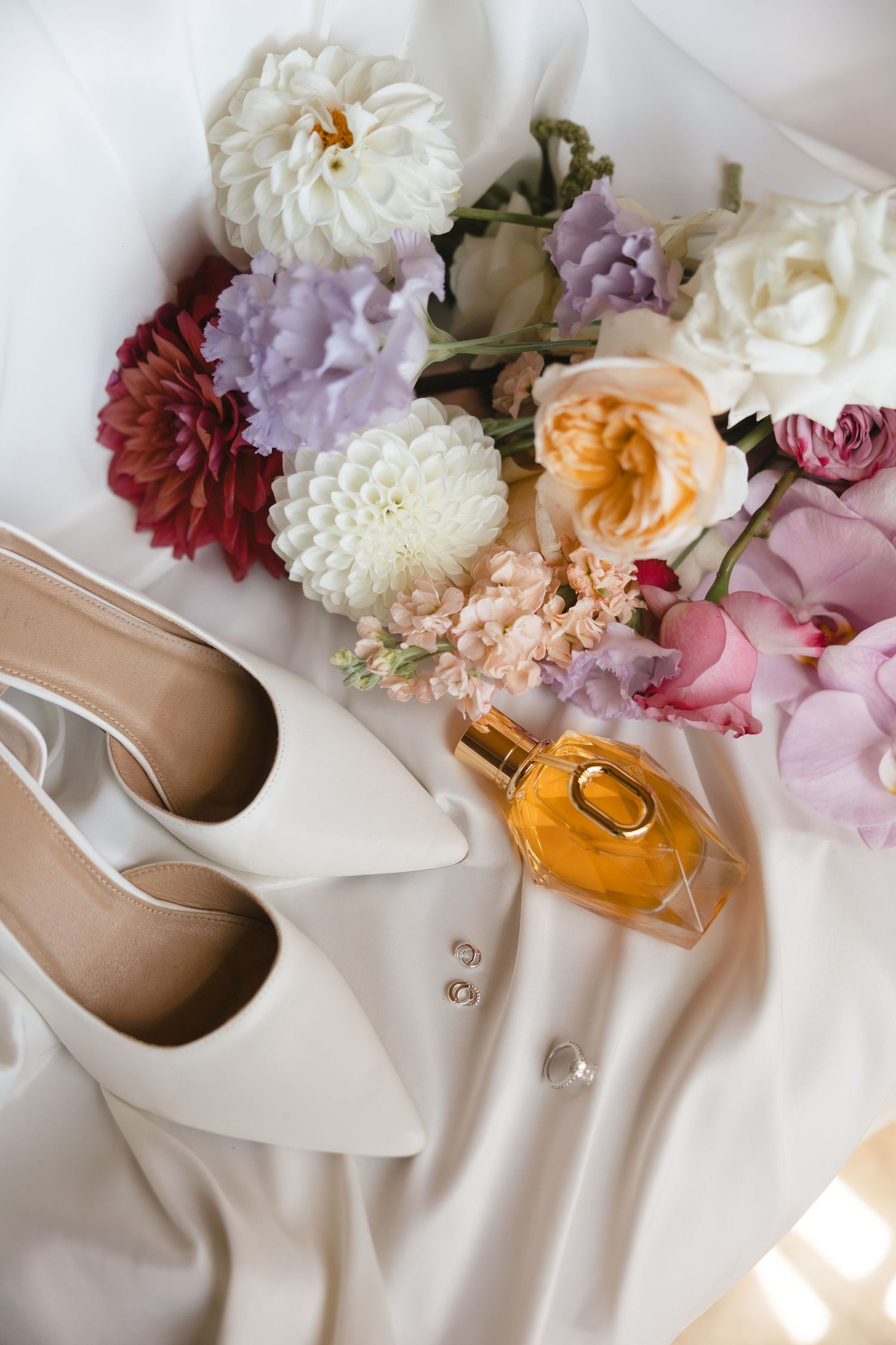 Wedding details featuring white heels, flowers and perfume arranged on silk fabric.