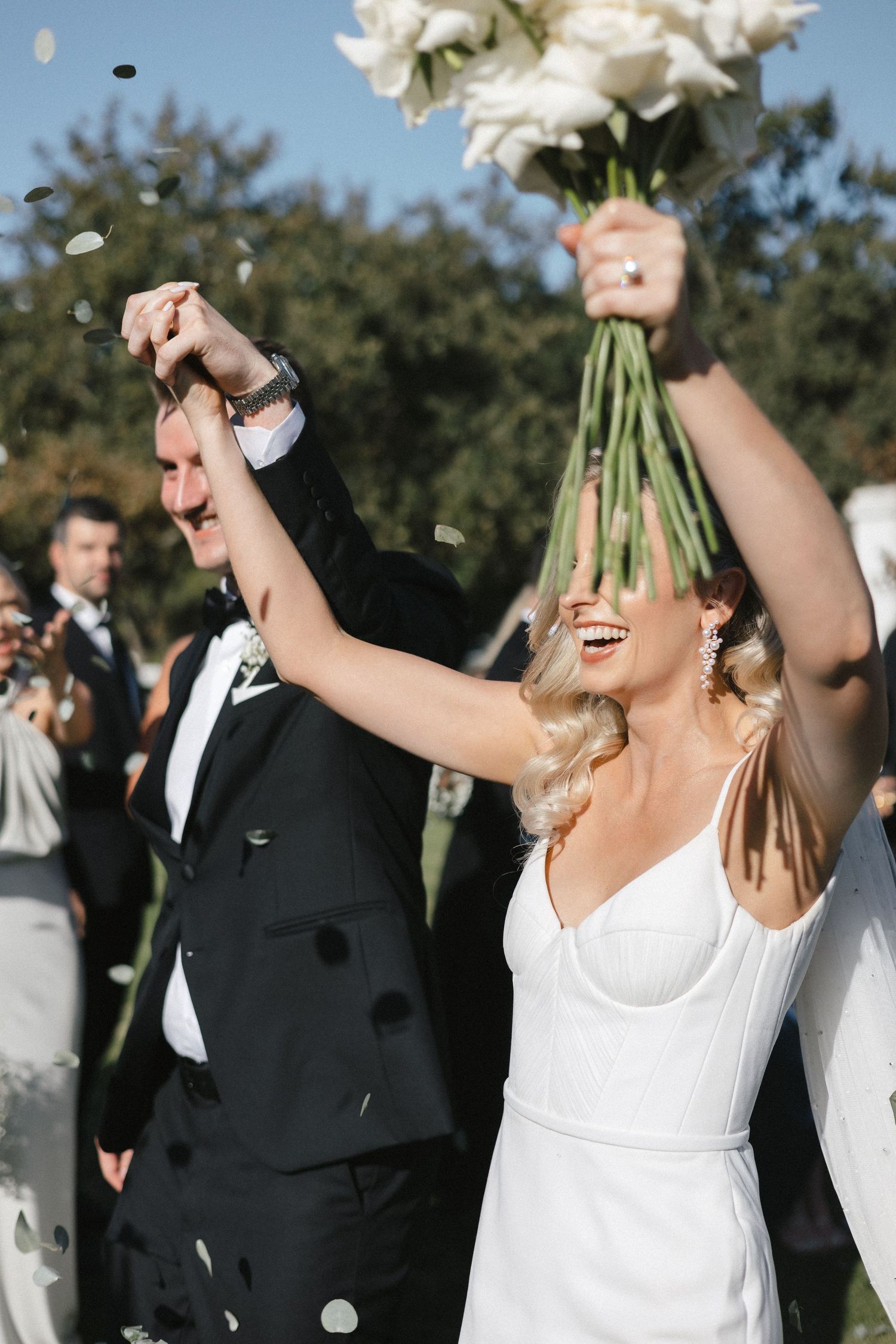 A joyful wedding celebration with raised hands and a white bouquet against an outdoor backdrop.
