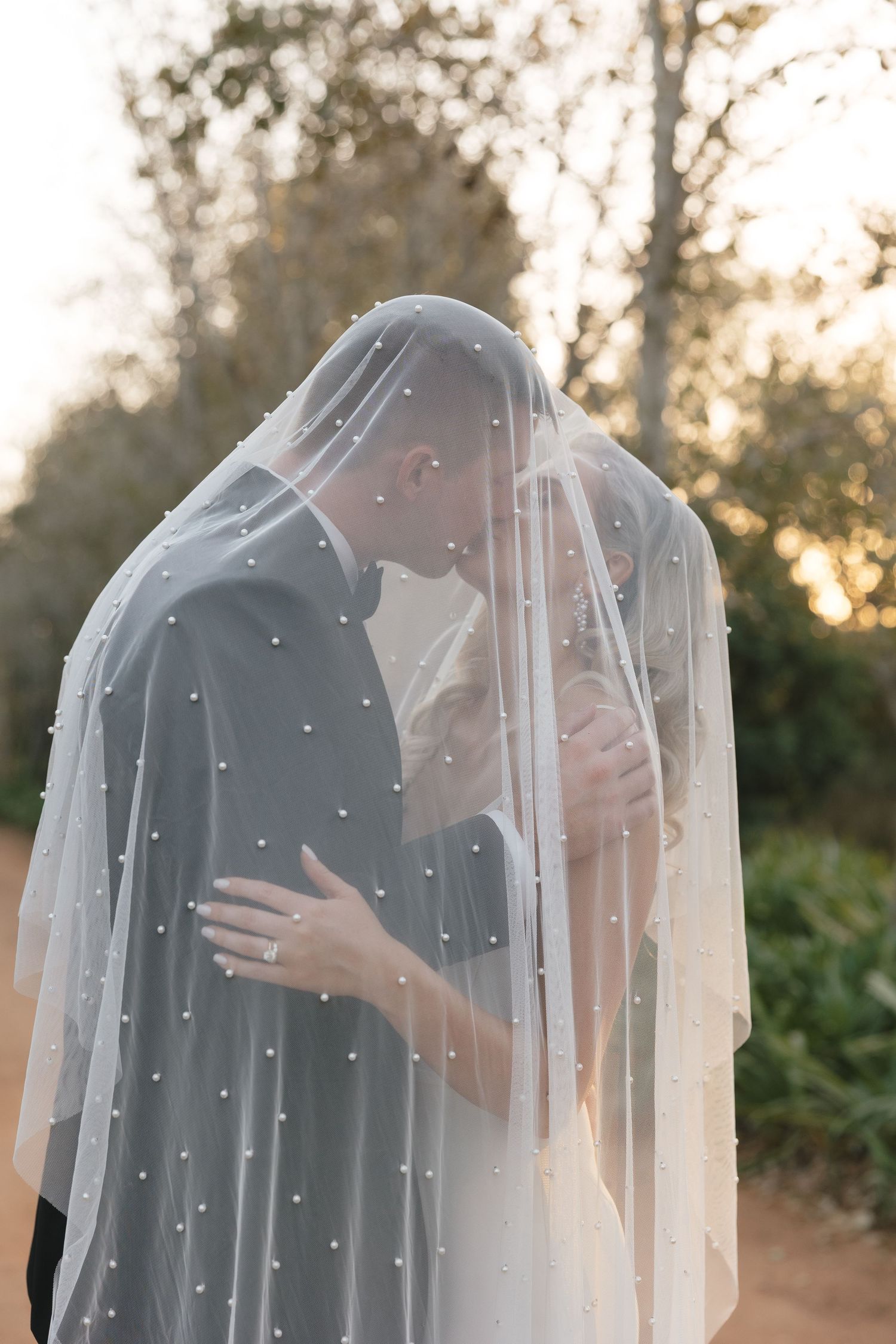 A romantic moment under a pearl-studded veil during sunset at an outdoor wedding.