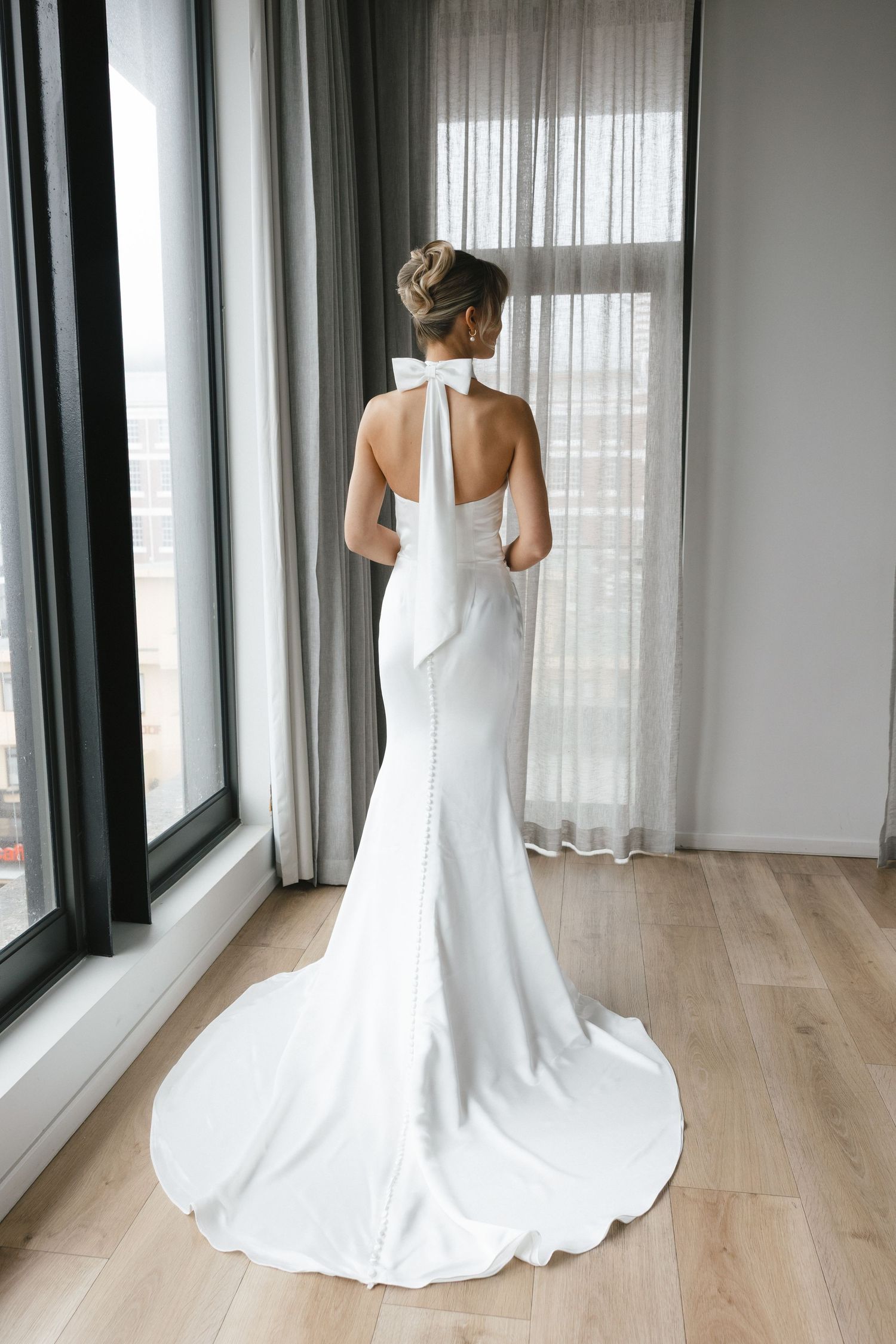 Full-length view of a sleek white wedding dress with train and halter neckline against bright windows.
