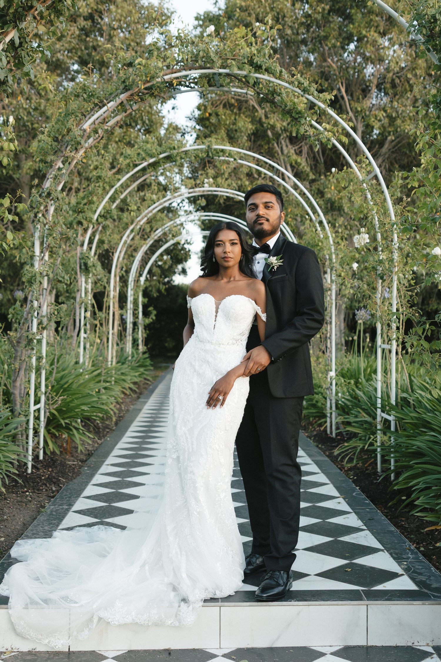 A couple stands under an arched garden walkway with checkerboard flooring.
