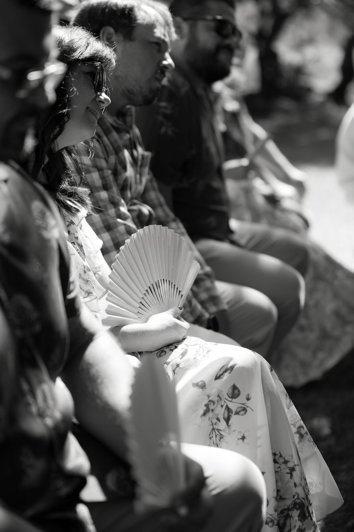 Black and white image of wedding guests holding decorative fans during an outdoor ceremony.