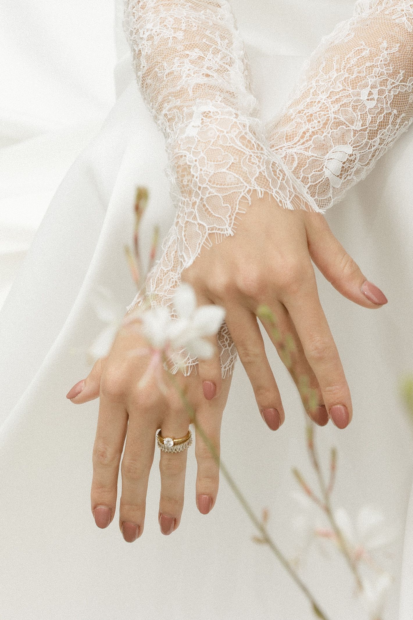 Close-up view of hands wearing delicate white lace gloves and an engagement ring against a light background.
