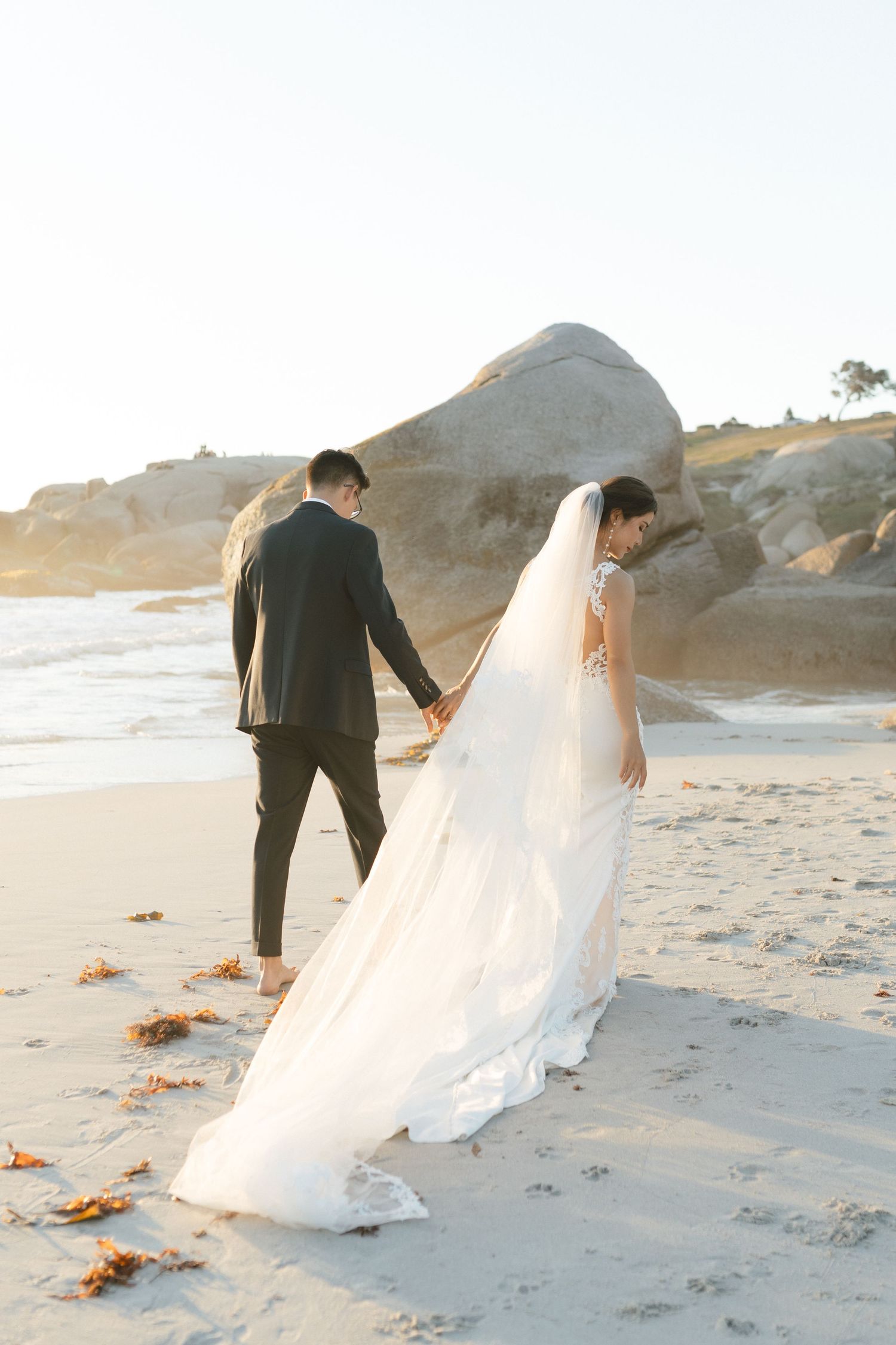 Couple walking on beach with flowing wedding dress and veil in golden light.