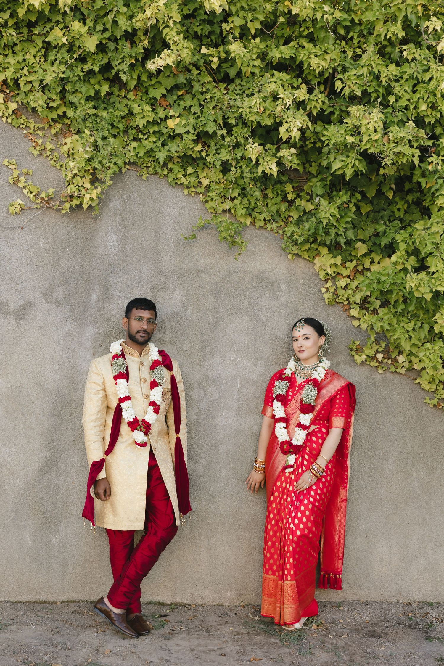 A couple in traditional Indian wedding attire poses against a grey wall with green ivy.