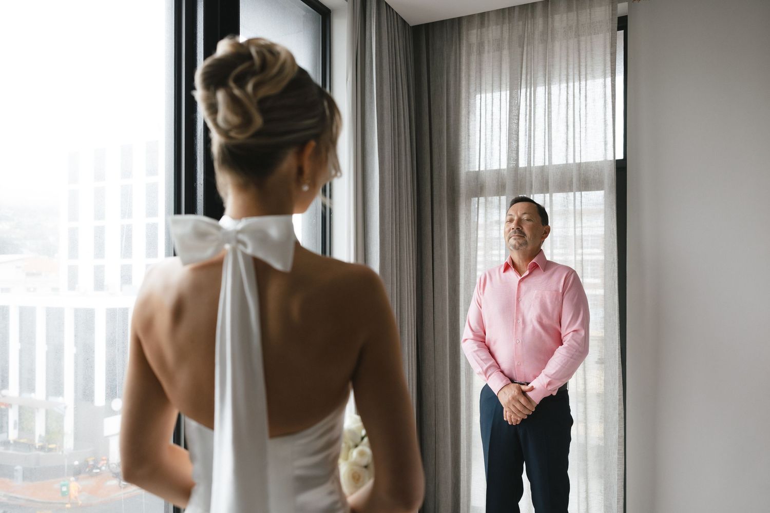 Wedding dress detail with bow neckline as figures stand by bright window.