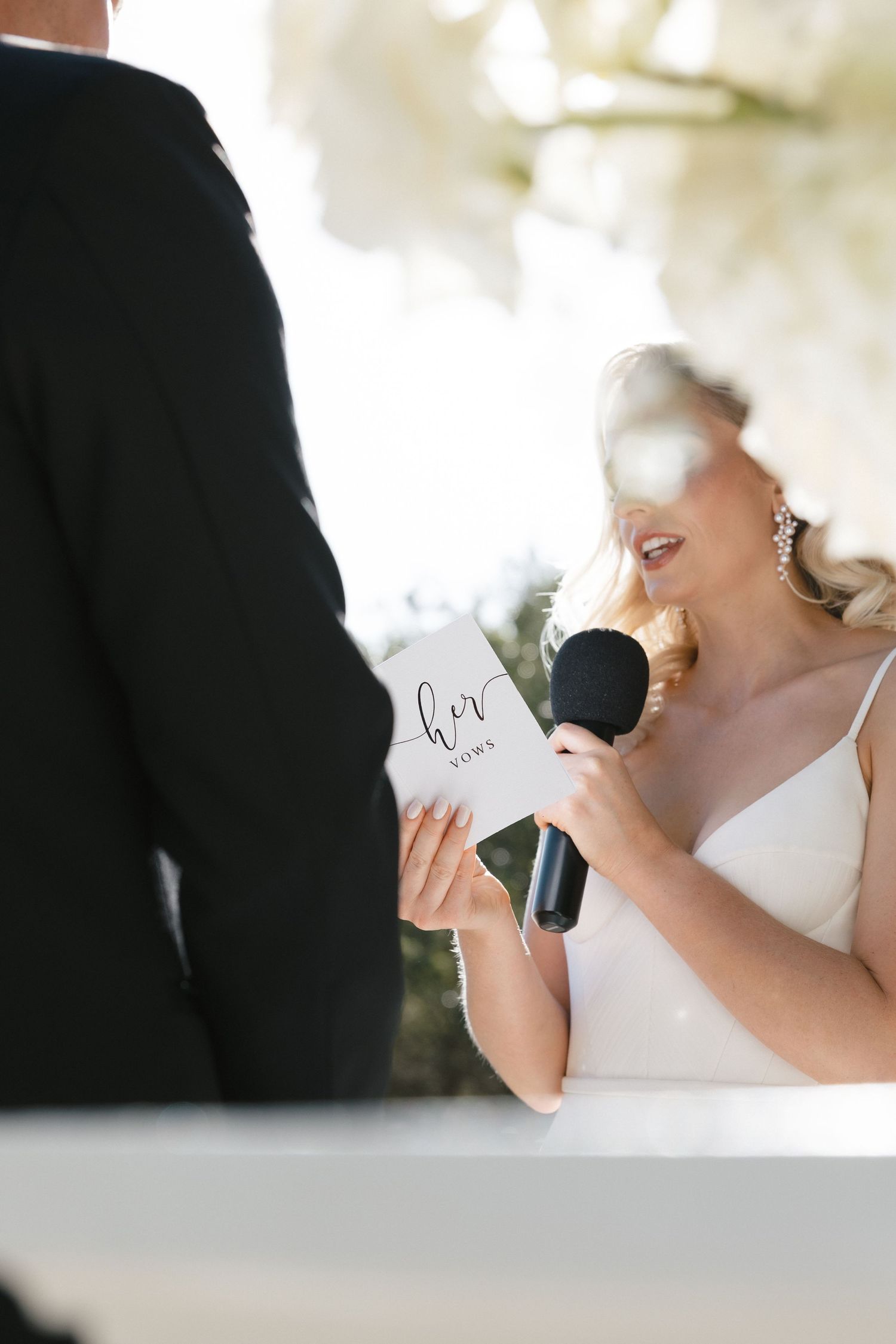 A wedding ceremony features the bride reading vows from a paper while holding a microphone.