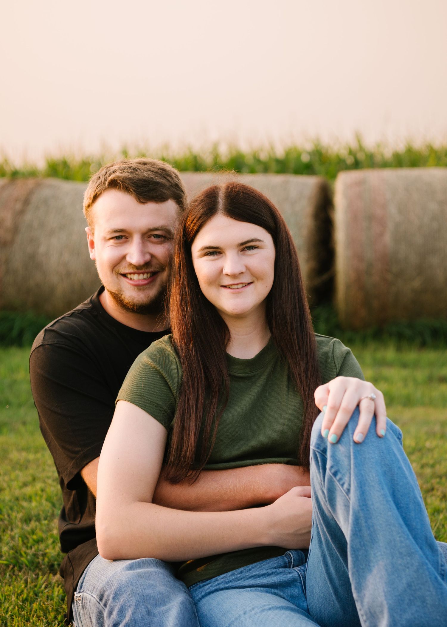 A couple sits together on grass with round hay bales in the background during a sunny outdoor photoshoot.