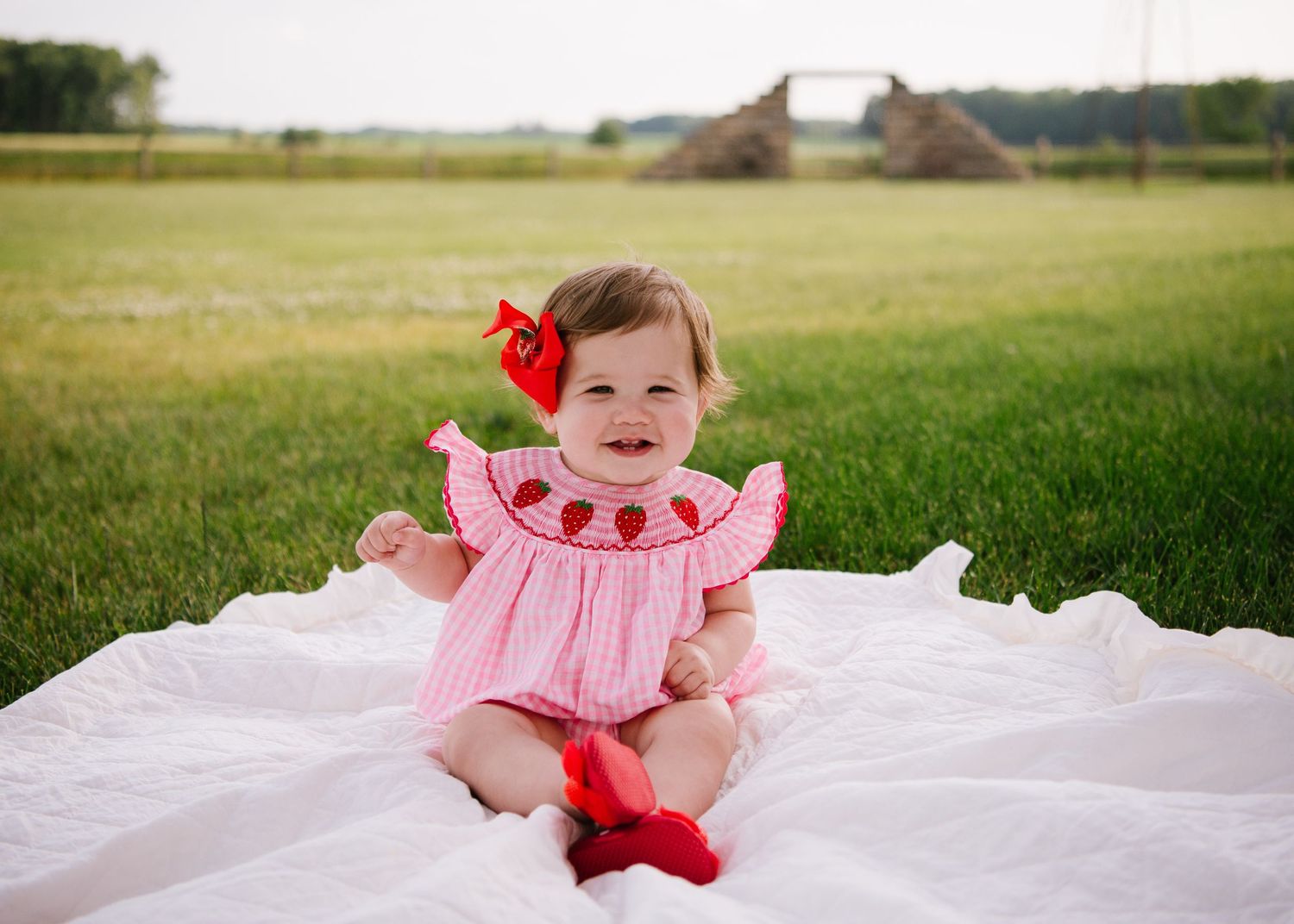 Baby wearing pink and red watermelon-themed outfit sits on white blanket in grassy field on sunny day.