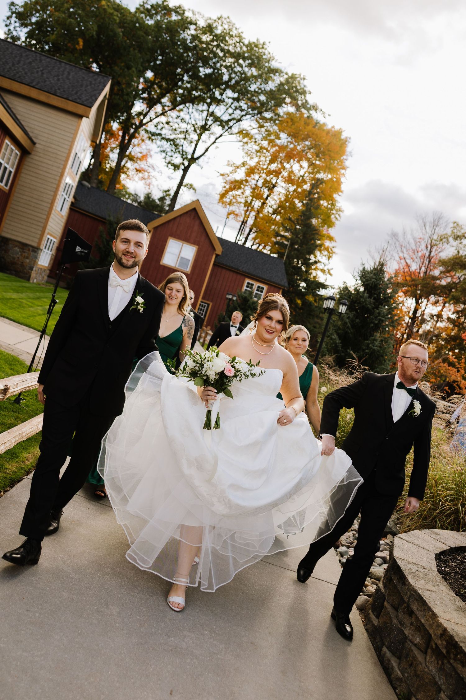Wedding party walks outdoors, bride in white gown holding bouquet, groom and groomsmen in black tuxedos, bridesmaids in green dresses.
