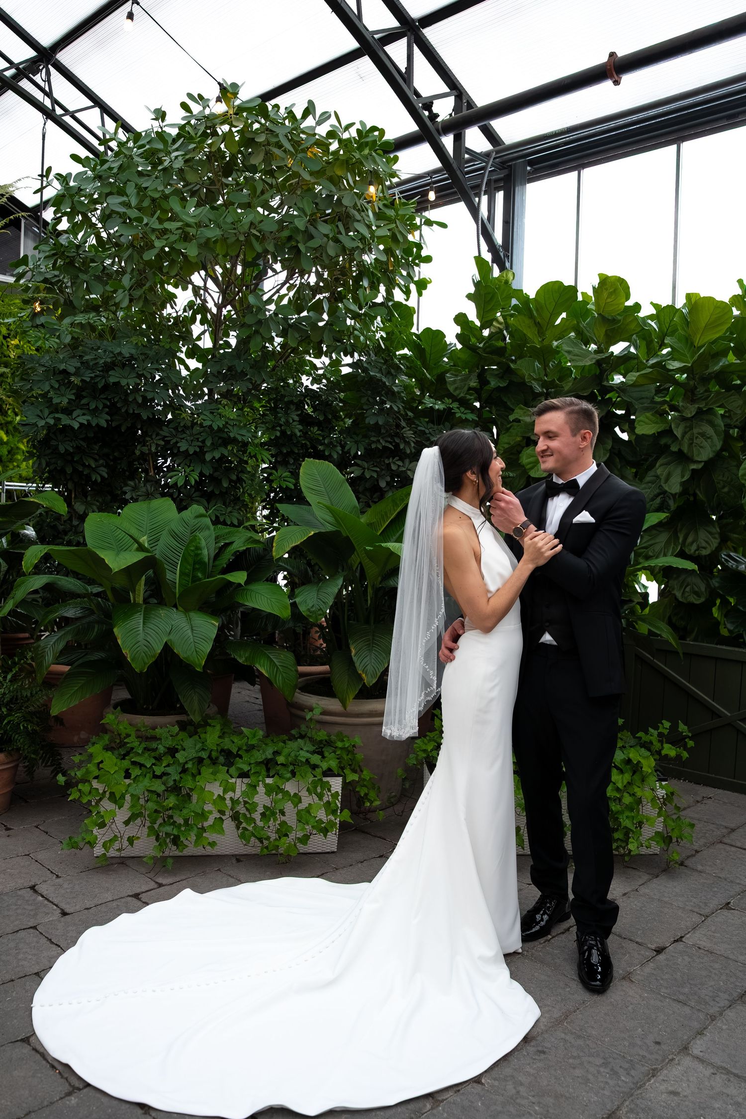 Bride in white gown with veil and groom in black tuxedo share a romantic moment in a lush greenhouse setting.