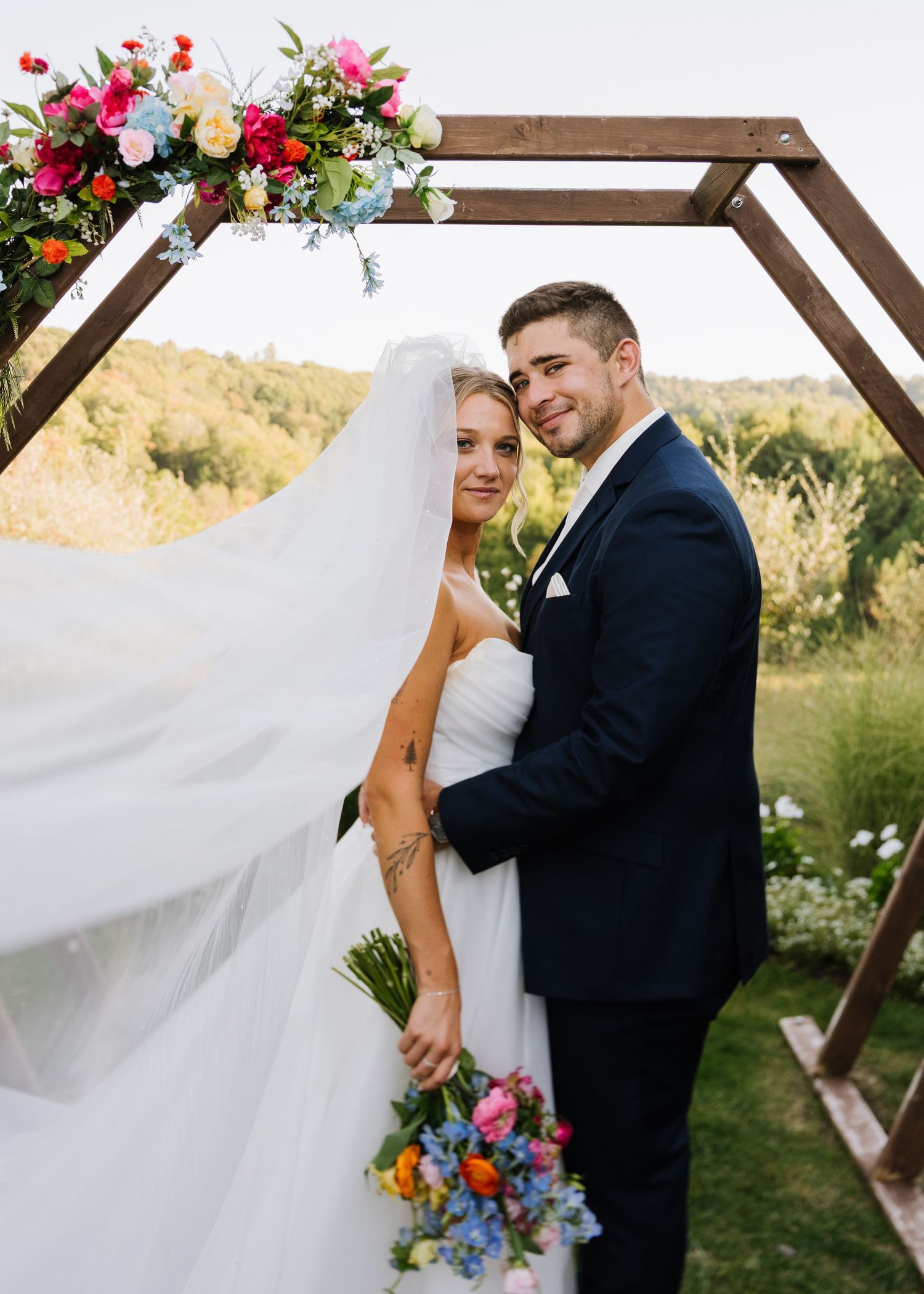 A romantic wedding couple embraces under a wooden arch decorated with vibrant pink and blue florals in an outdoor garden setting.
