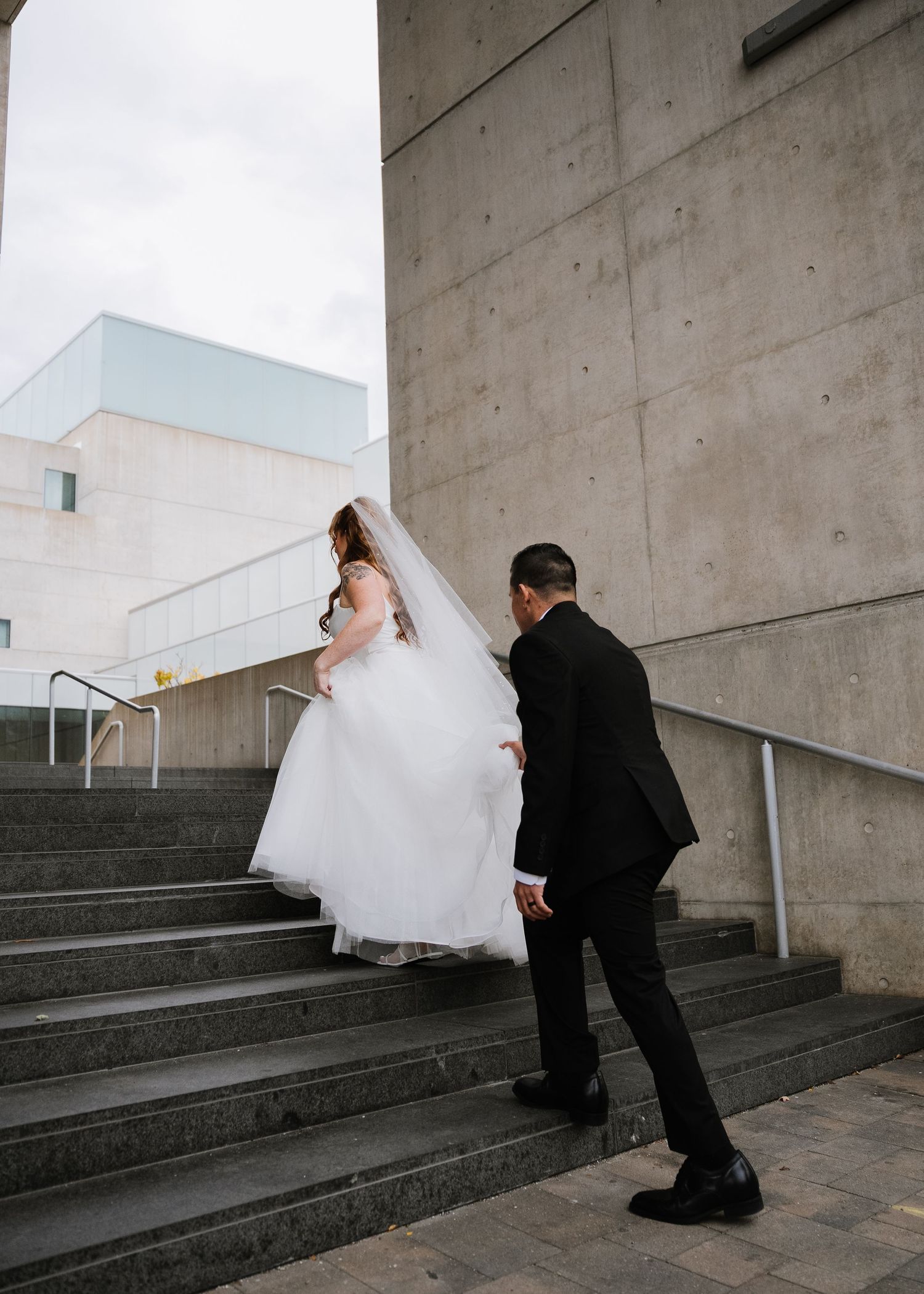 A bride in a white gown and veil walks up concrete steps with a groom in a black suit at an urban venue.