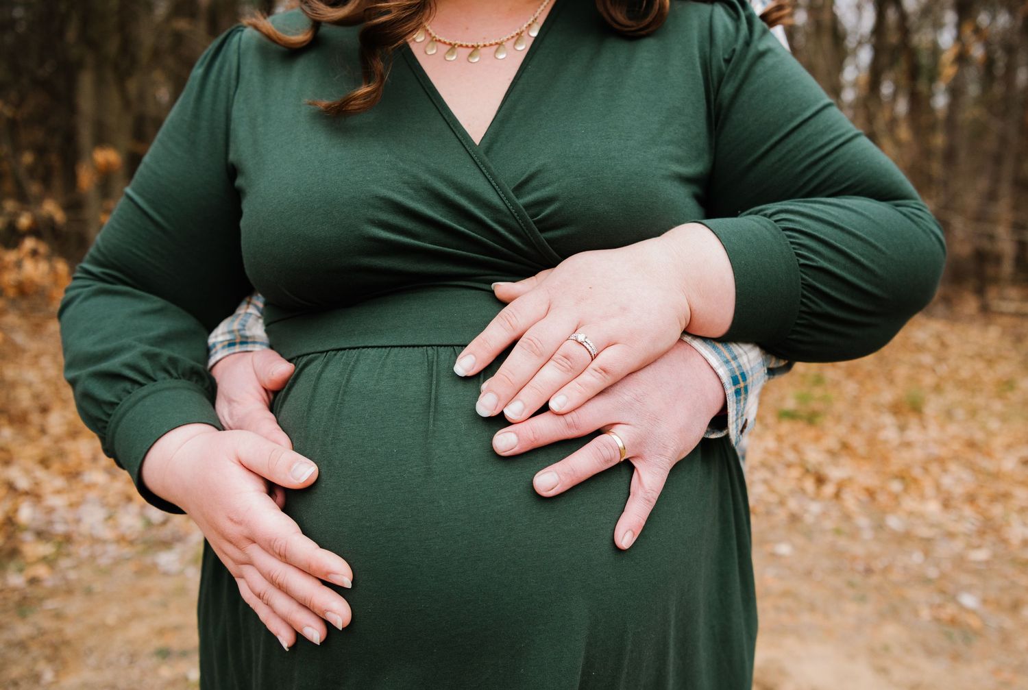 Hands gently resting on a baby bump, wearing an emerald green wrap dress against an autumn forest backdrop.