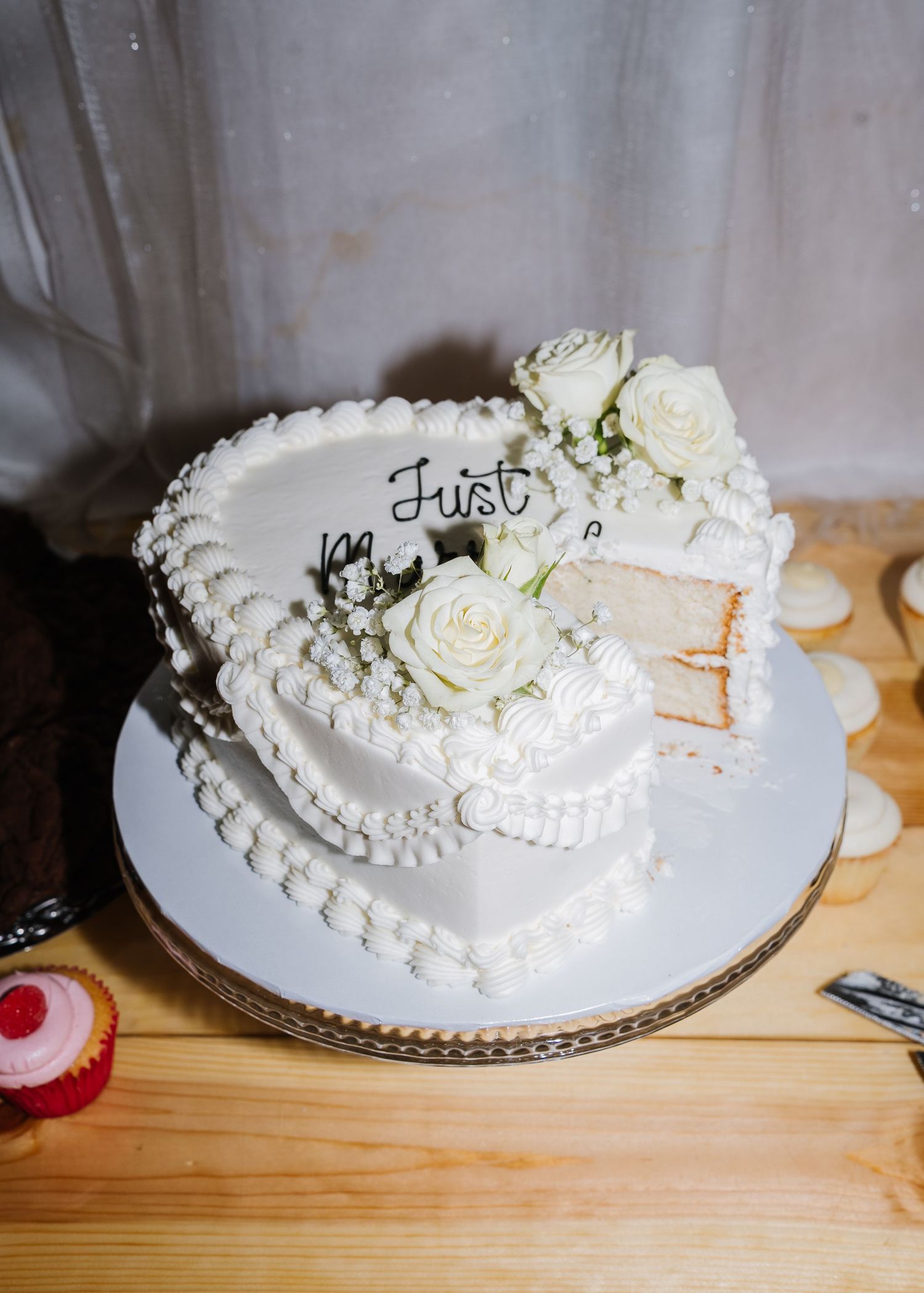 A white two-tiered wedding cake decorated with white roses and piped details sits on a wooden surface.