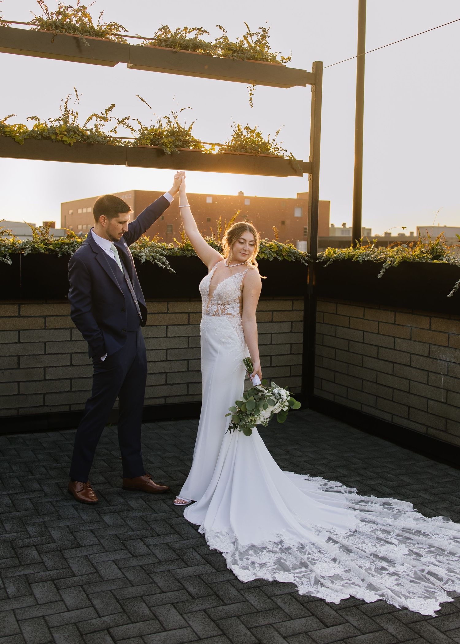 Newlywed couple celebrates on rooftop terrace with long white wedding dress and bouquet at golden hour sunset.