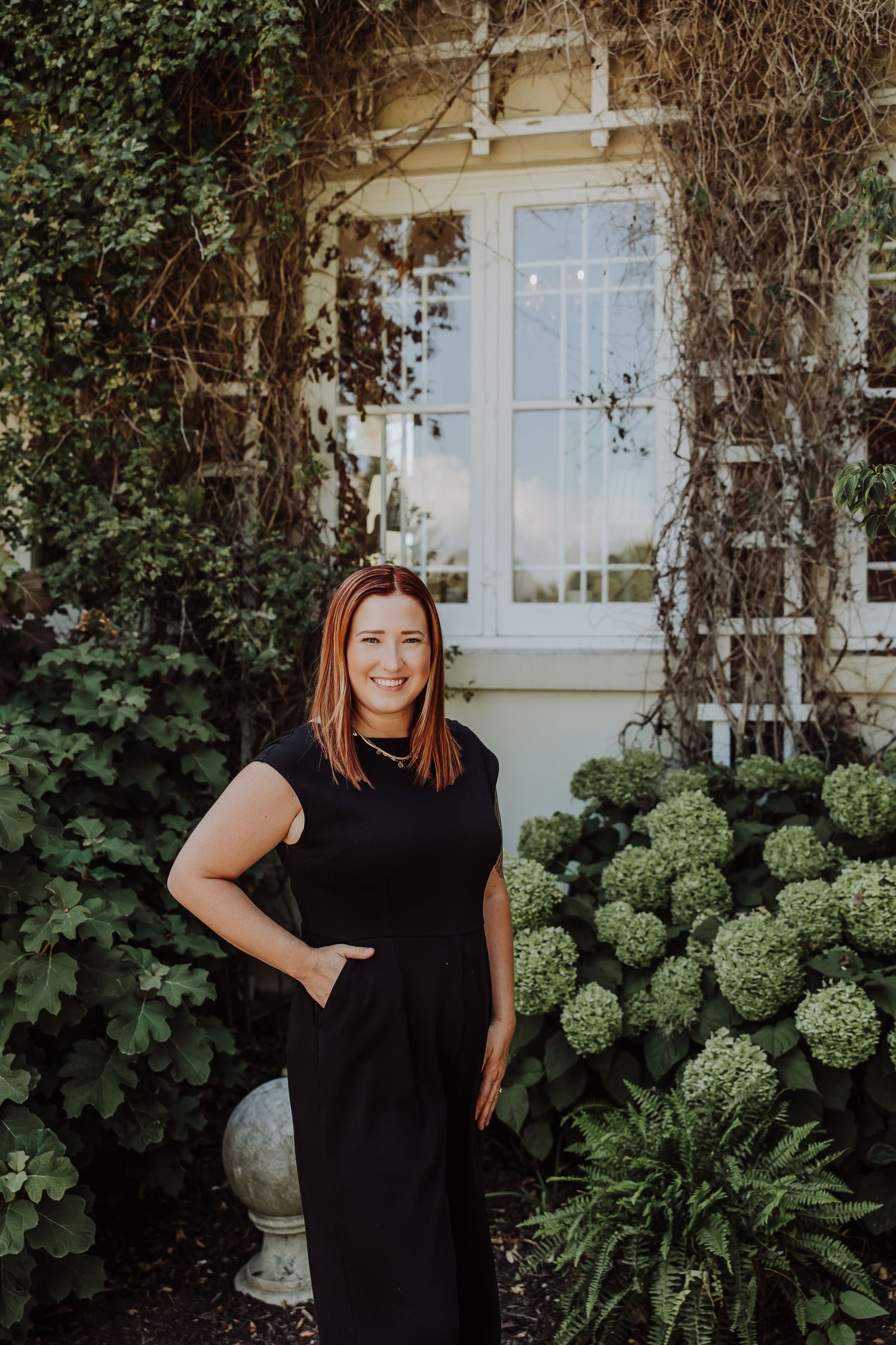 Woman in black dress smiling in lush garden with hydrangeas and ivy-covered wall behind her.