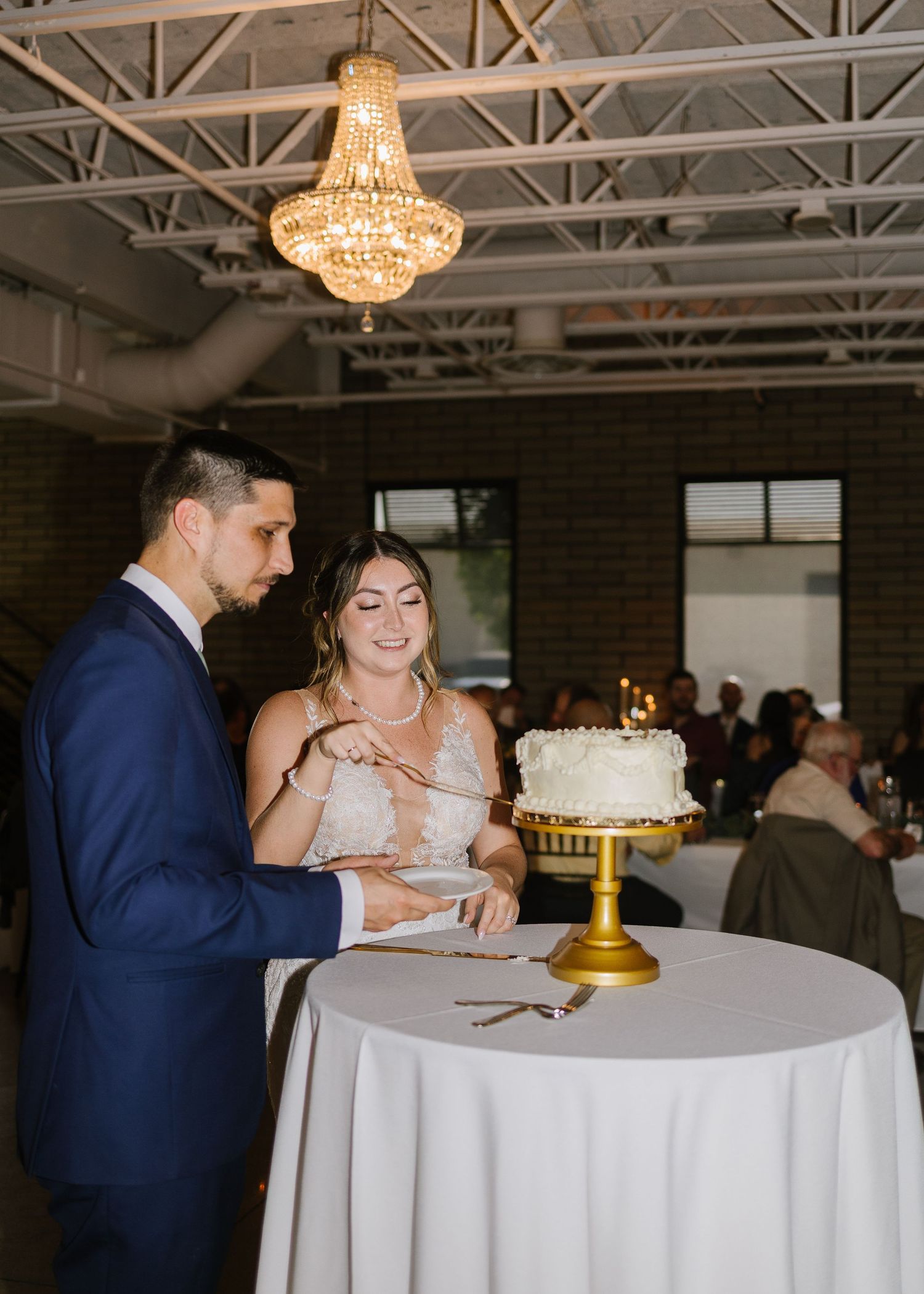 An elegant couple cutting their wedding cake together under a crystal chandelier at an industrial-style venue.