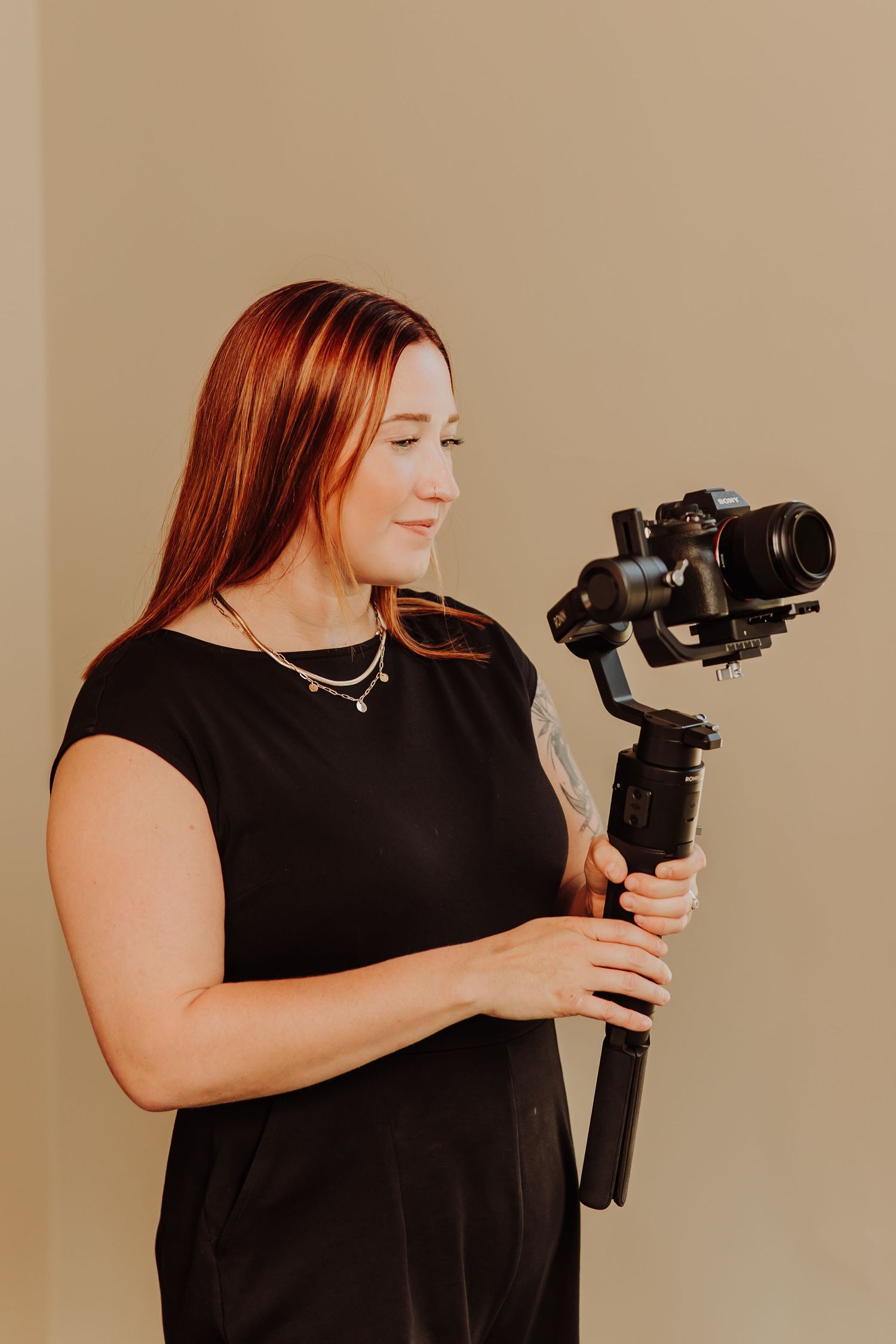 Woman in black dress holding a camera gimbal stabilizer, smiling softly against a neutral beige background.