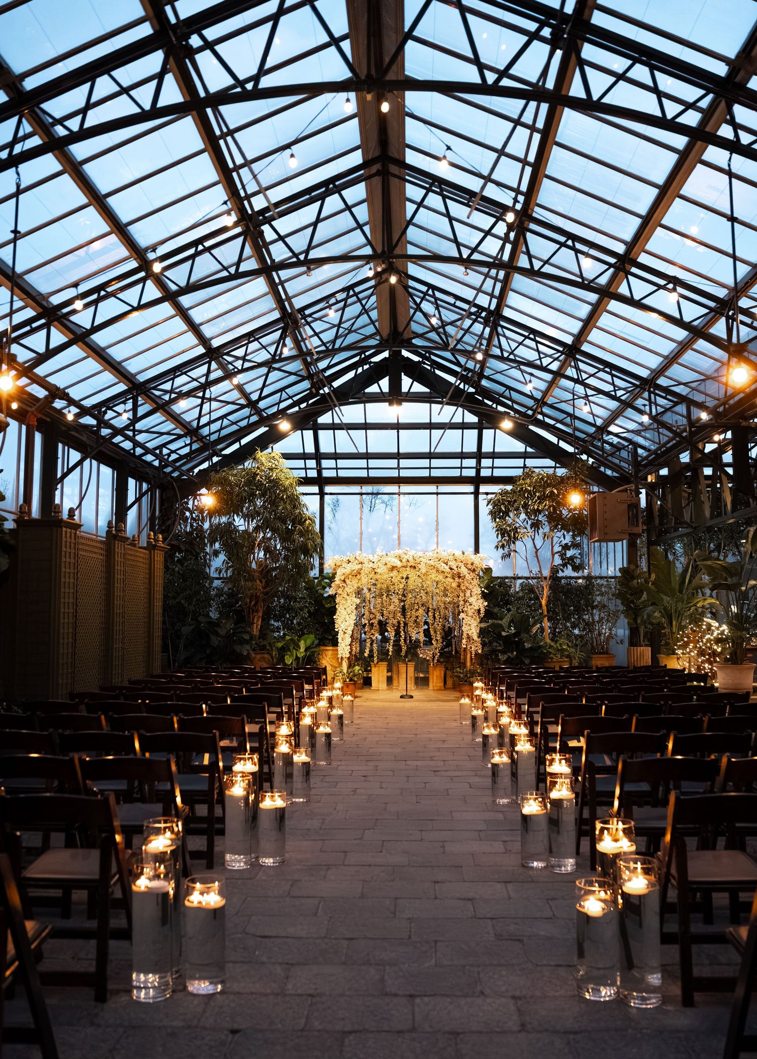 Elegant indoor wedding ceremony in a glass greenhouse with candlelit aisle and white floral arch altar.