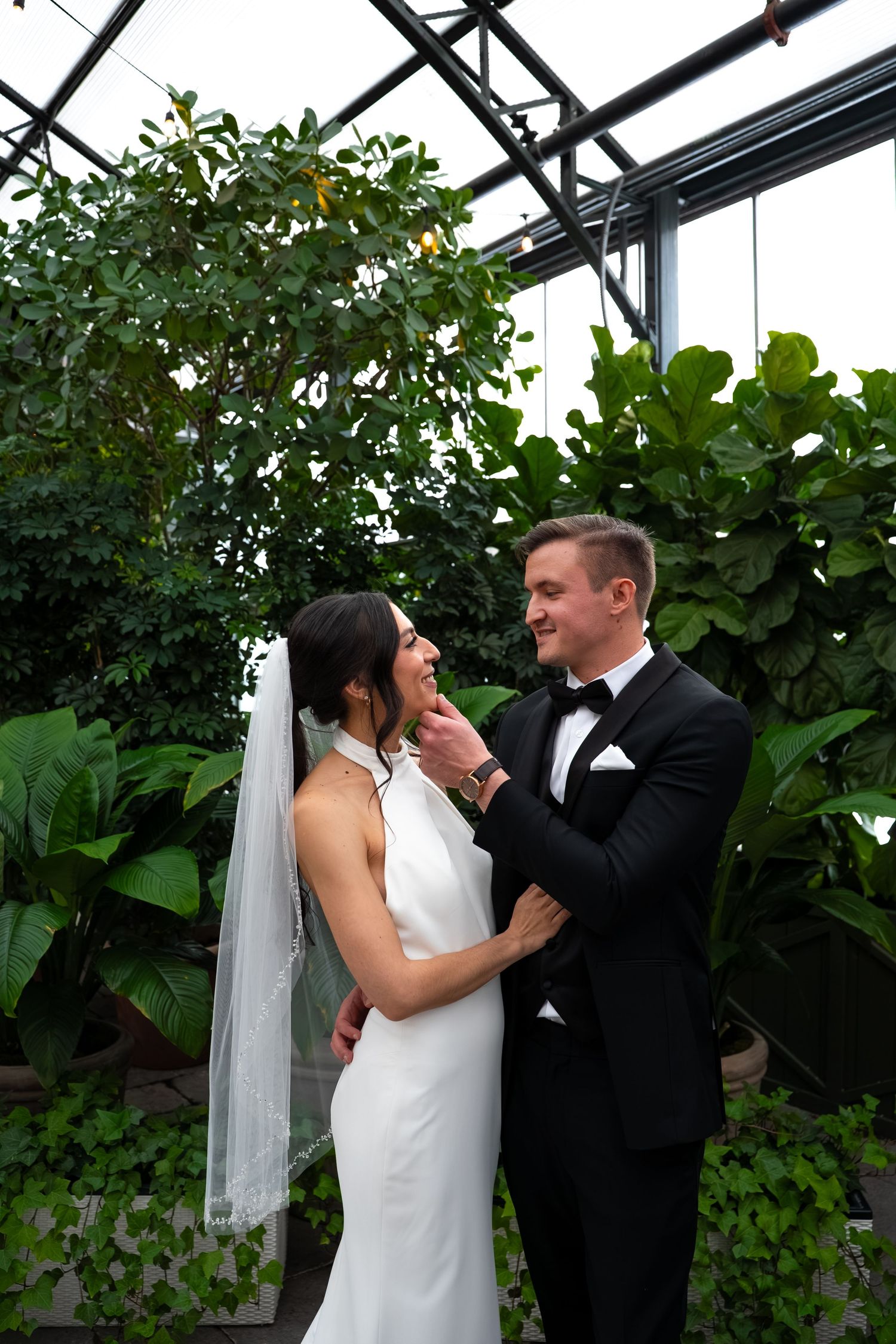 A bride and groom share a romantic moment in a lush greenhouse, with the bride in a white gown and veil.