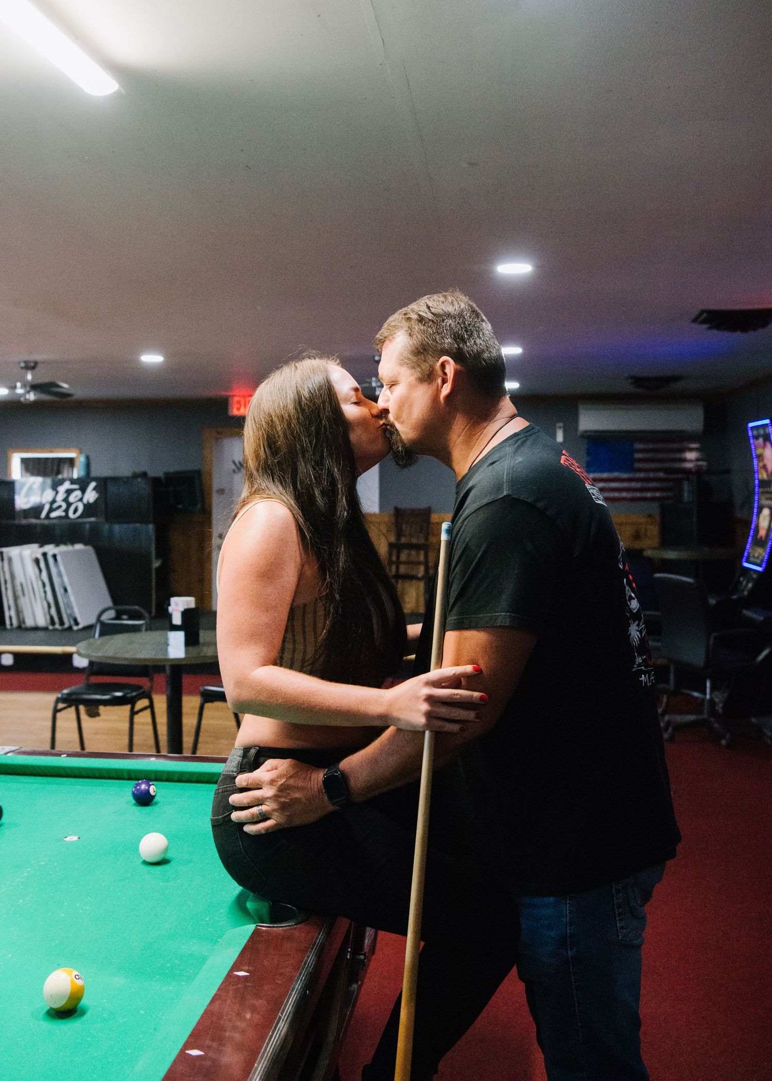 A romantic couple shares an intimate moment while playing pool in a dimly lit game room with arcade machines and an American flag.