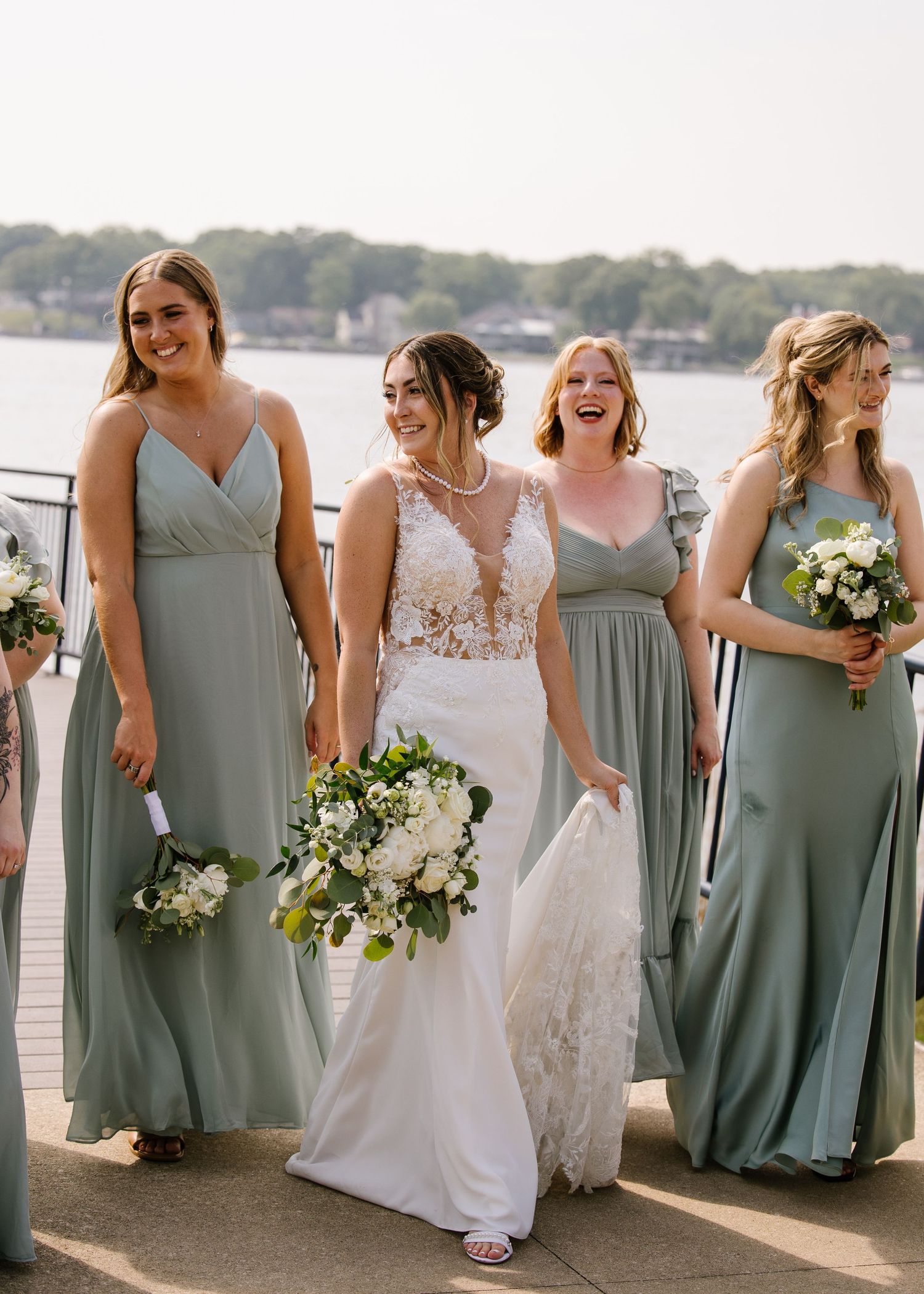 A wedding party in matching sage green dresses and a white bridal gown pose together by the waterfront.