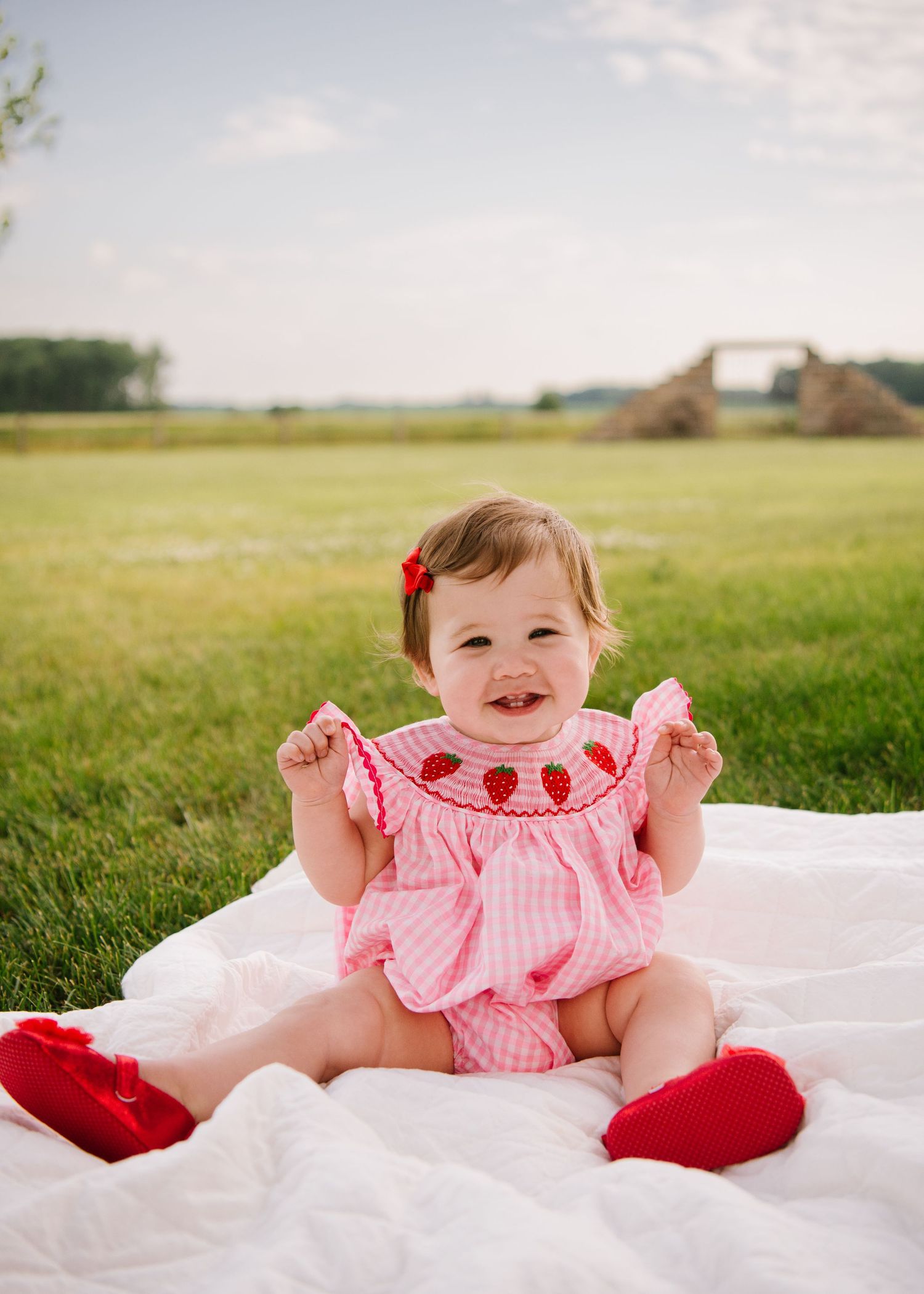 Happy baby wearing pink ruffled outfit laying on white blanket in grassy field with red accessories.
