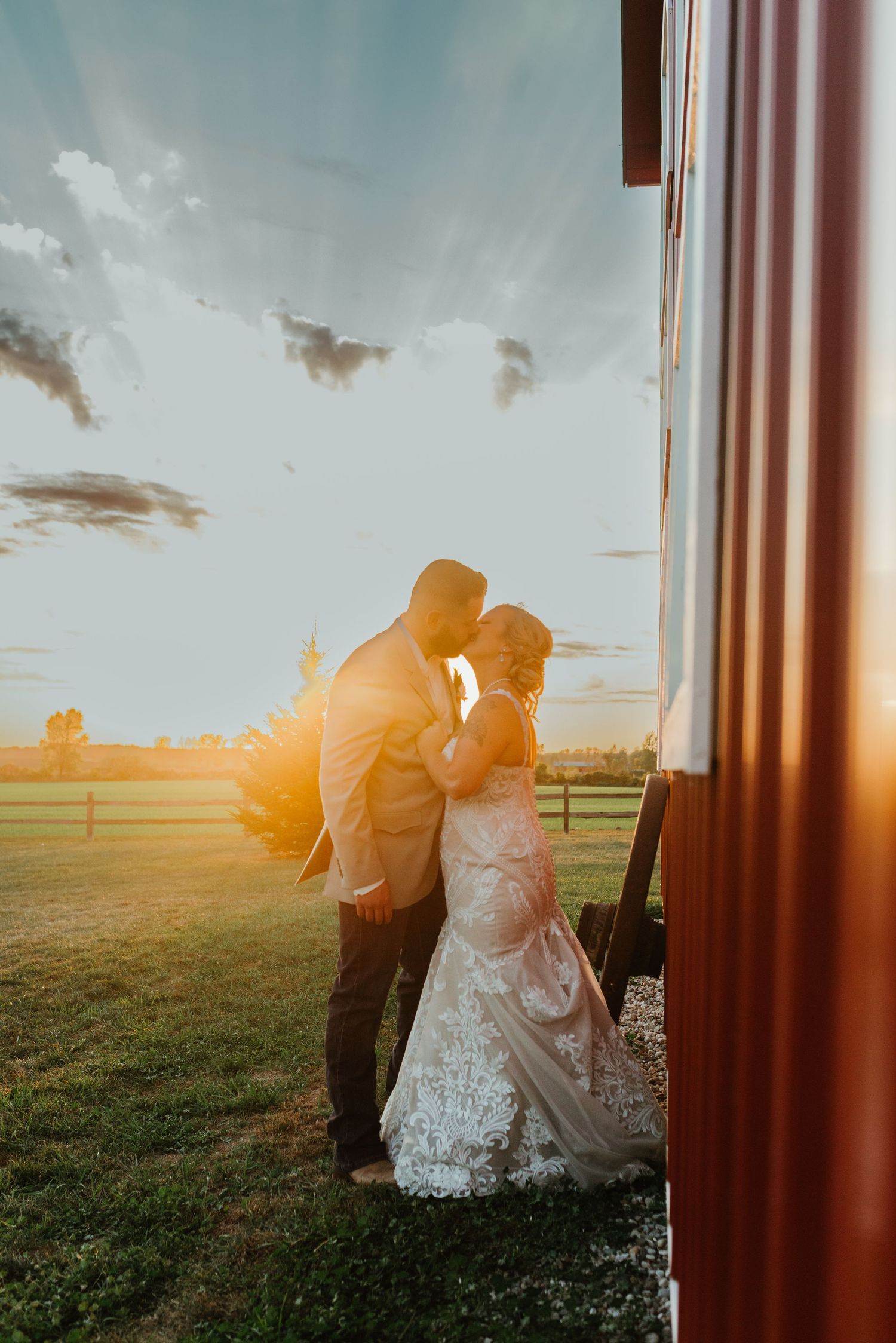 A romantic wedding couple embraces at sunset, with golden light streaming through a rustic barn doorway setting.