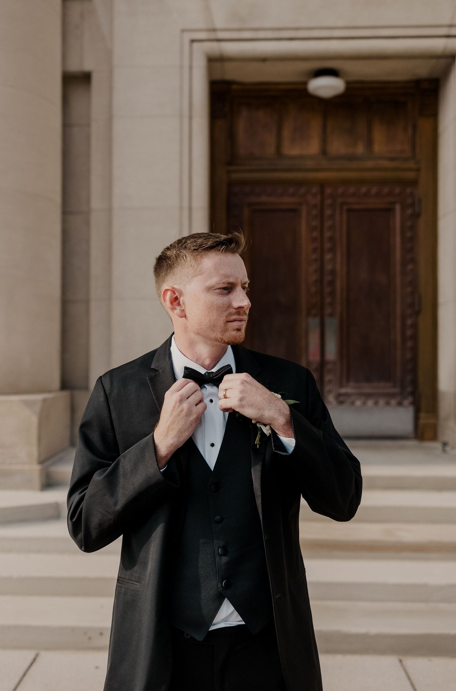 Someone adjusts their bowtie while wearing a classic black tuxedo outside an ornate building entrance.