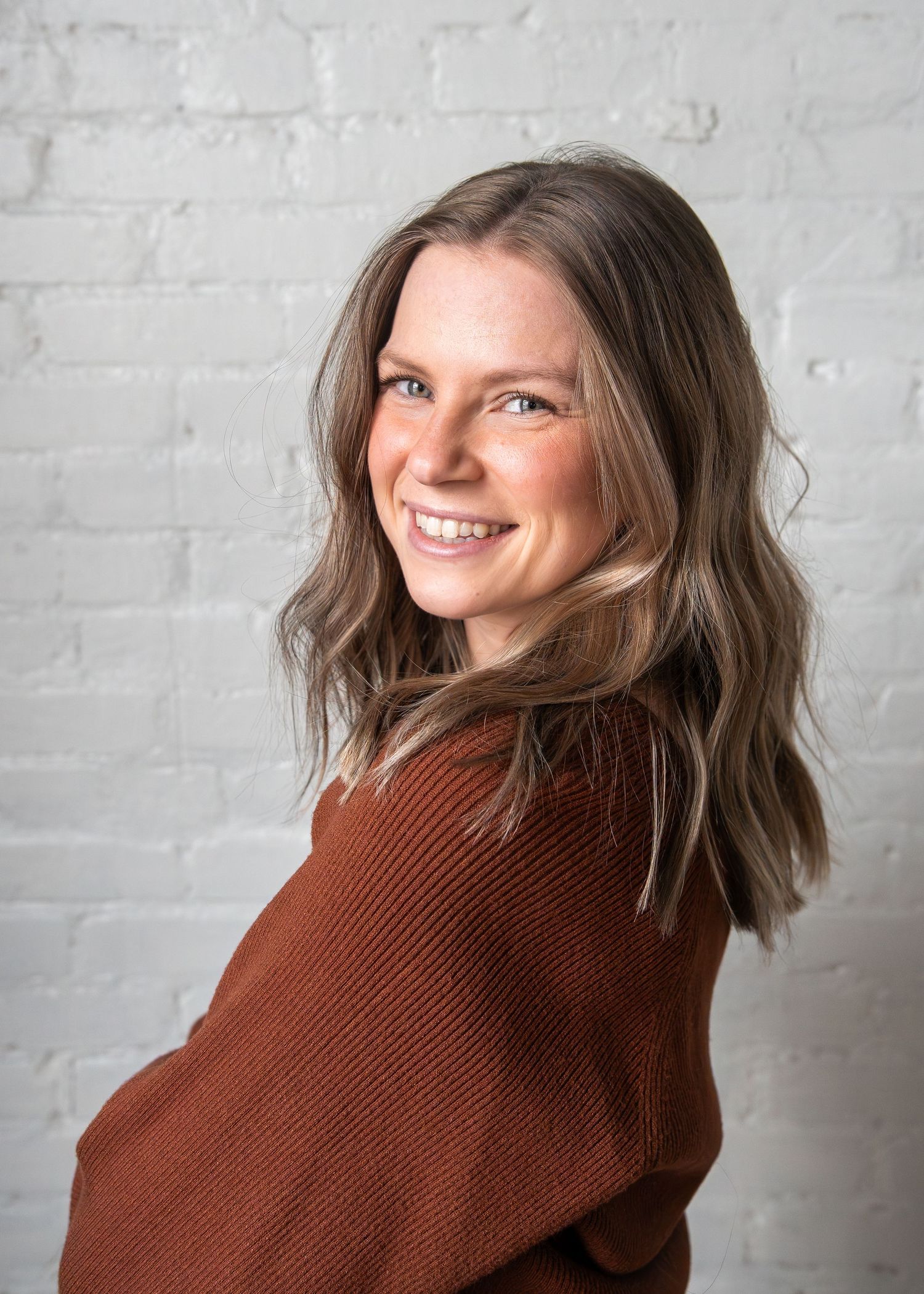 Smiling woman with brown hair wearing a rust sweater, posing against a white brick wall.