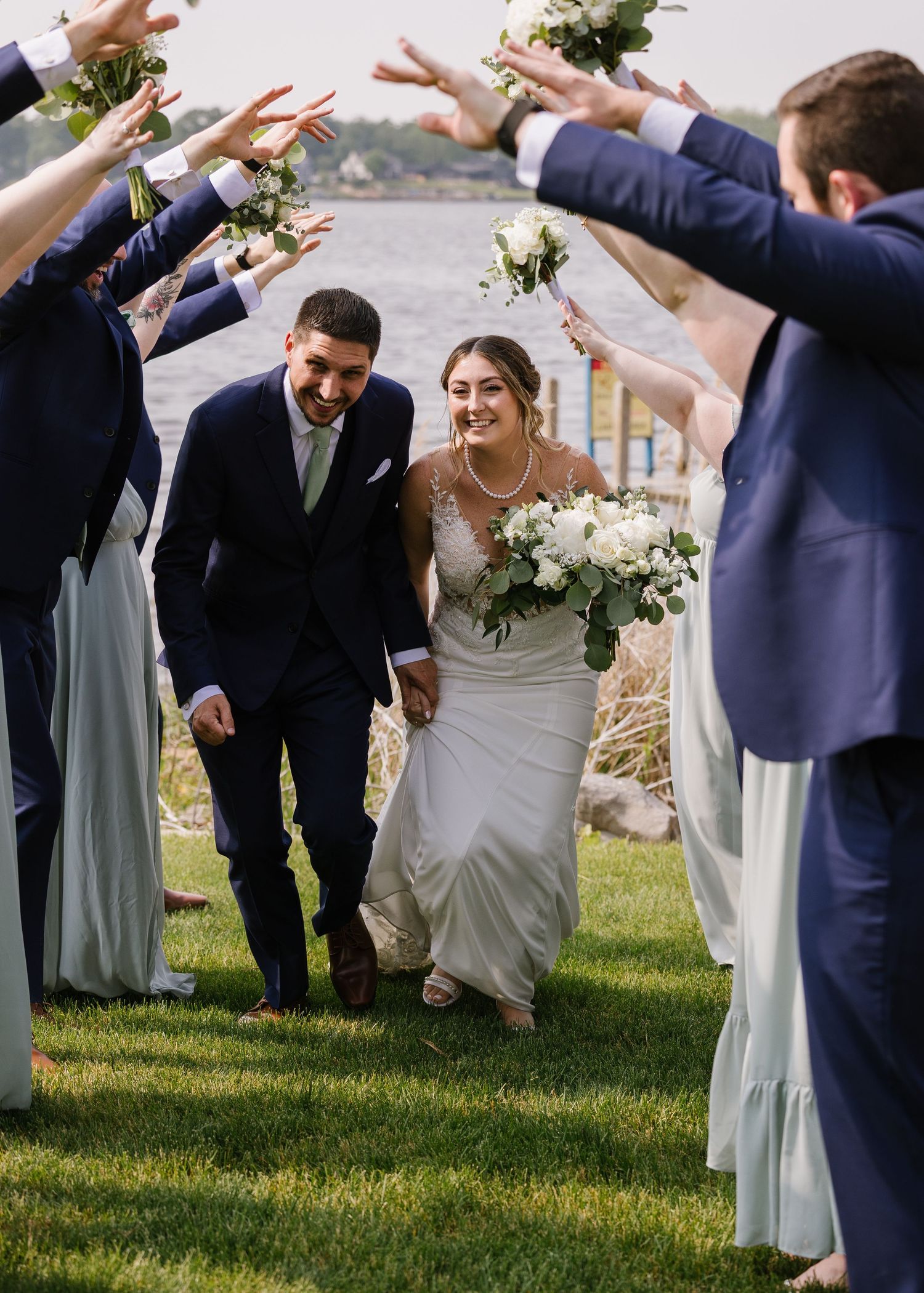 Wedding couple walks through an outdoor aisle lined with friends in blue dresses holding white bouquets over their heads.