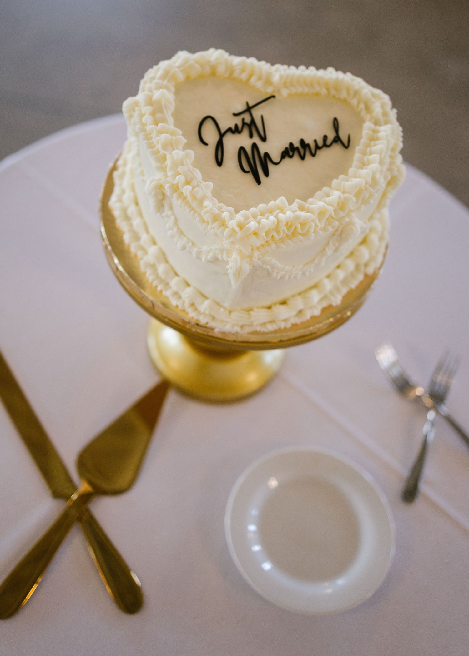 Heart-shaped white wedding cake with 'Just Married' text displayed on gold cake stand with cutlery on white tablecloth.