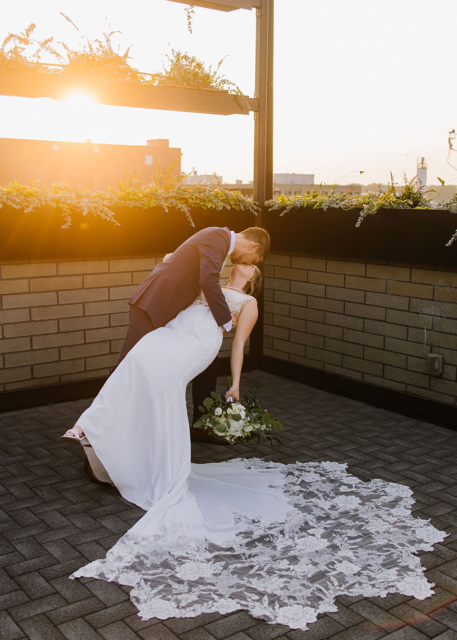 A couple shares a romantic moment at sunset on a rooftop terrace with a flowing white wedding dress trailing behind.