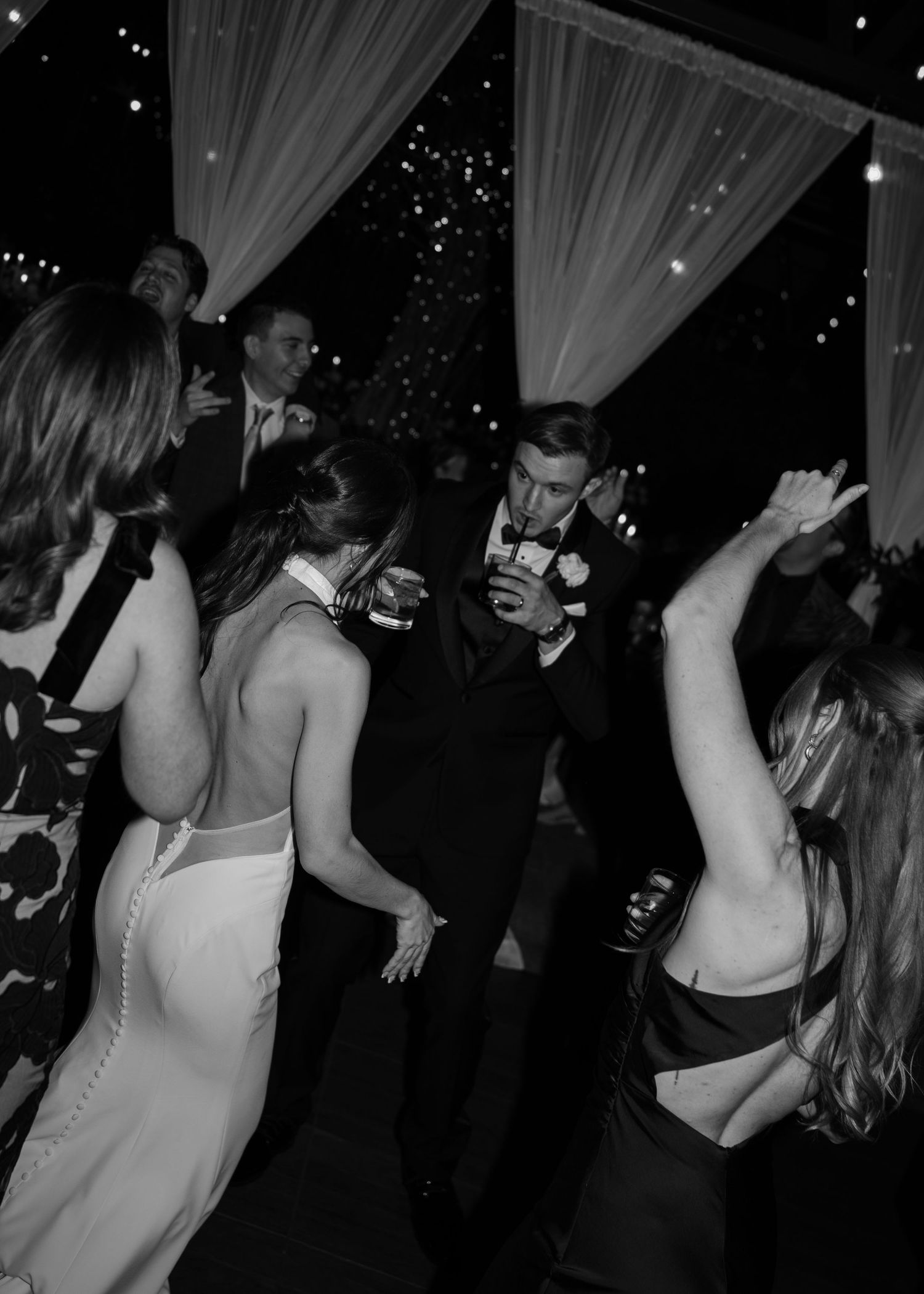 Black and white photo of wedding guests dancing joyfully at a elegant evening reception under draped fabric canopy.