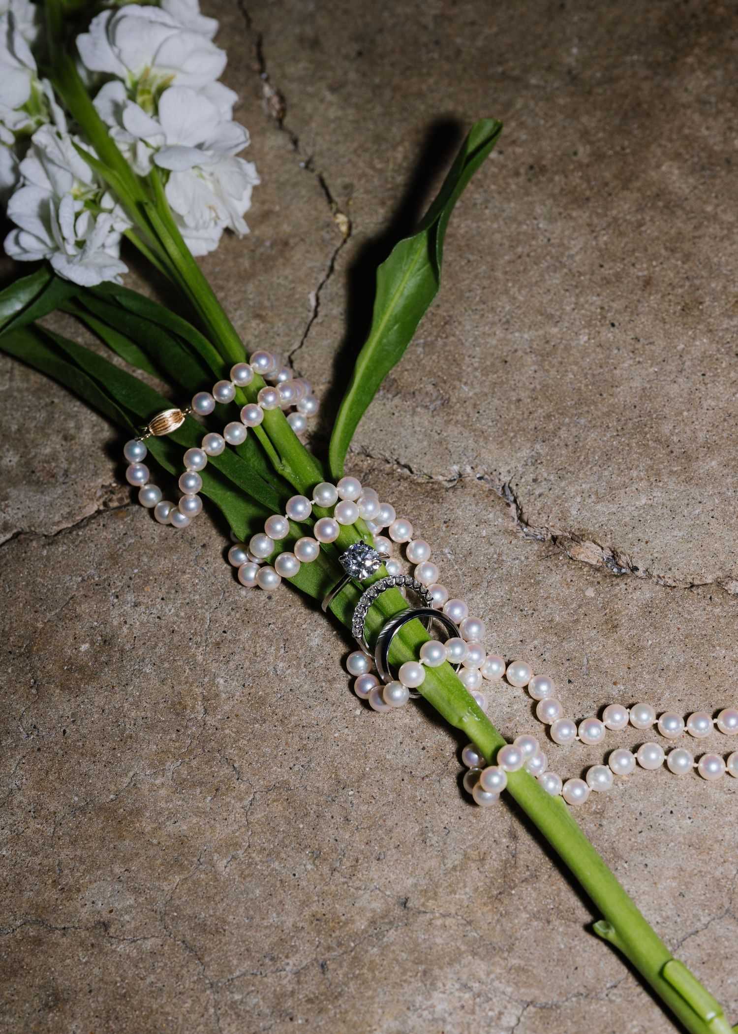 Pearl necklace draped around a green flower bouquet with white blooms on a stone surface.
