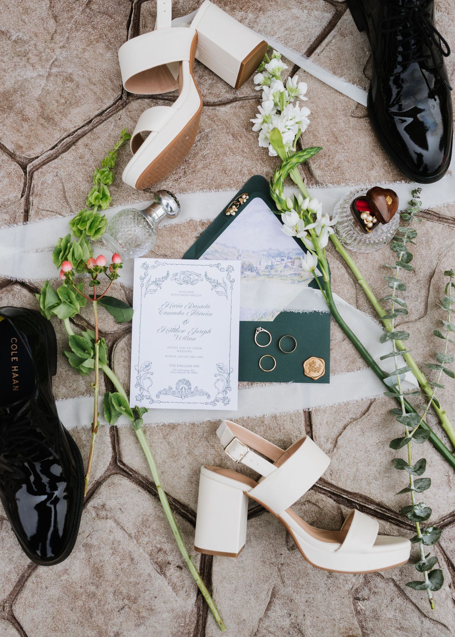 Wedding details flatlay with elegant invitation, rings on green velvet box, black dress shoes and white heels with floral accents.
